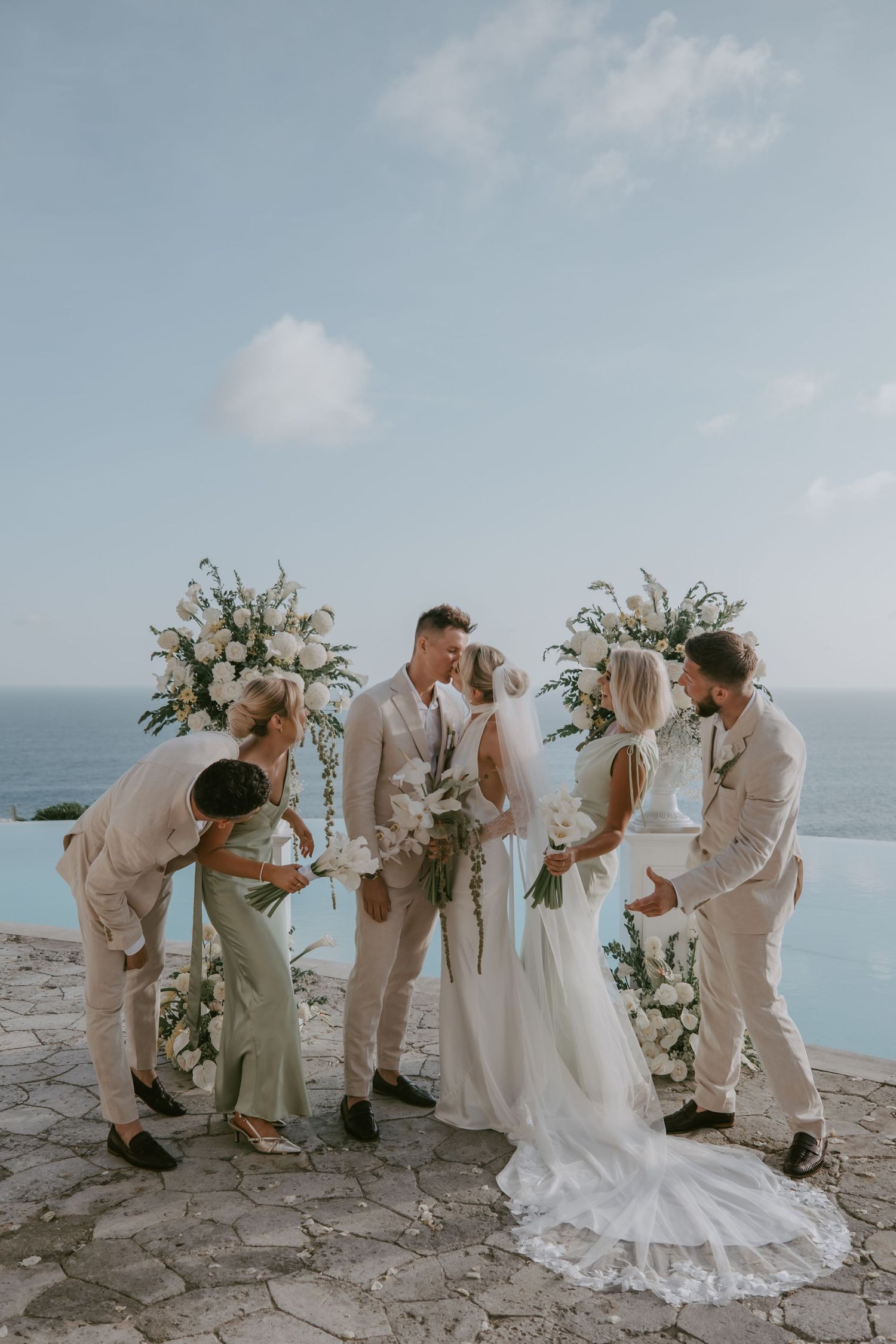 Wedding party celebrates with champagne on a stone terrace overlooking turquoise waters.