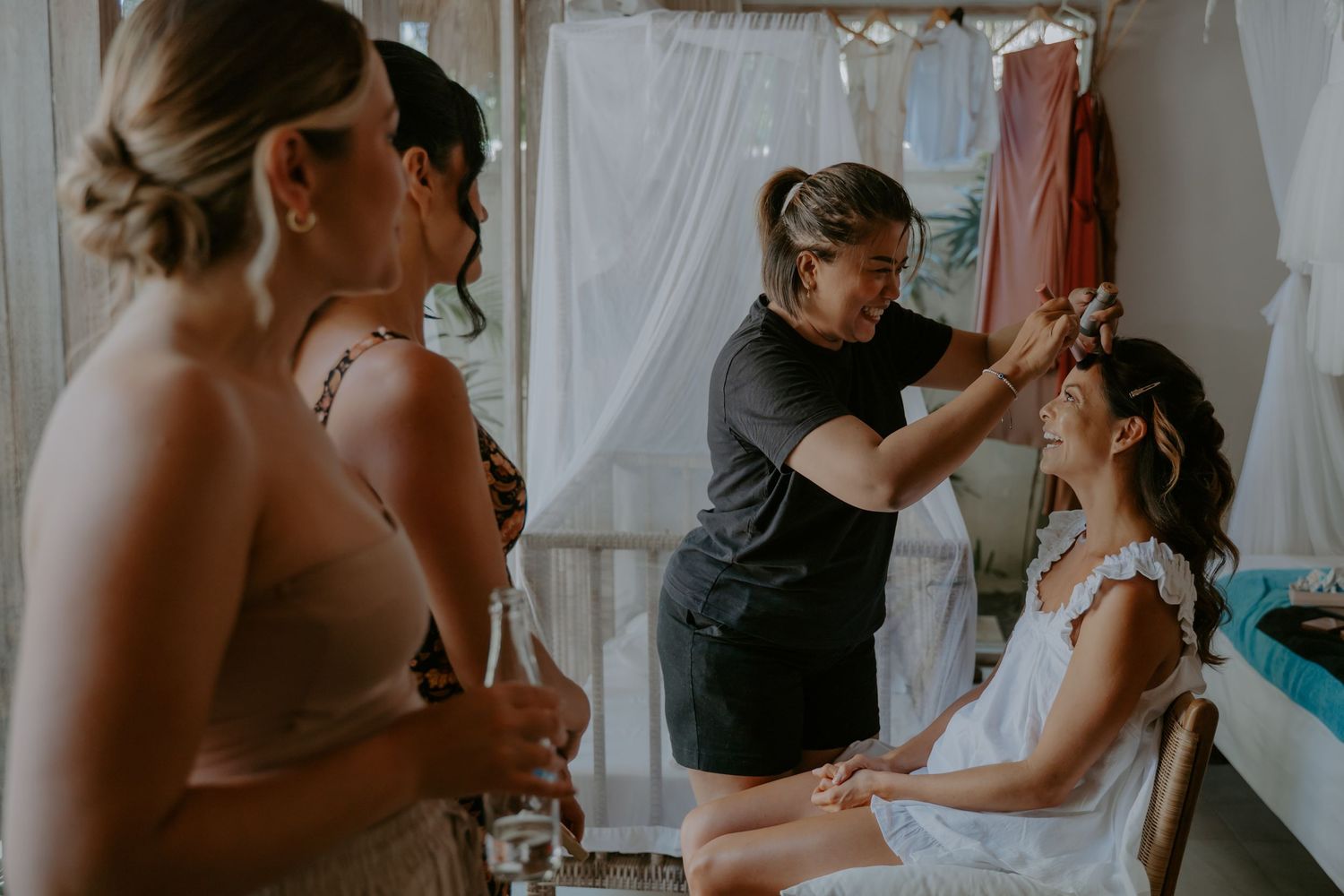 Wedding preparations underway as bridesmaids help with final touches in a sunlit room with white curtains.