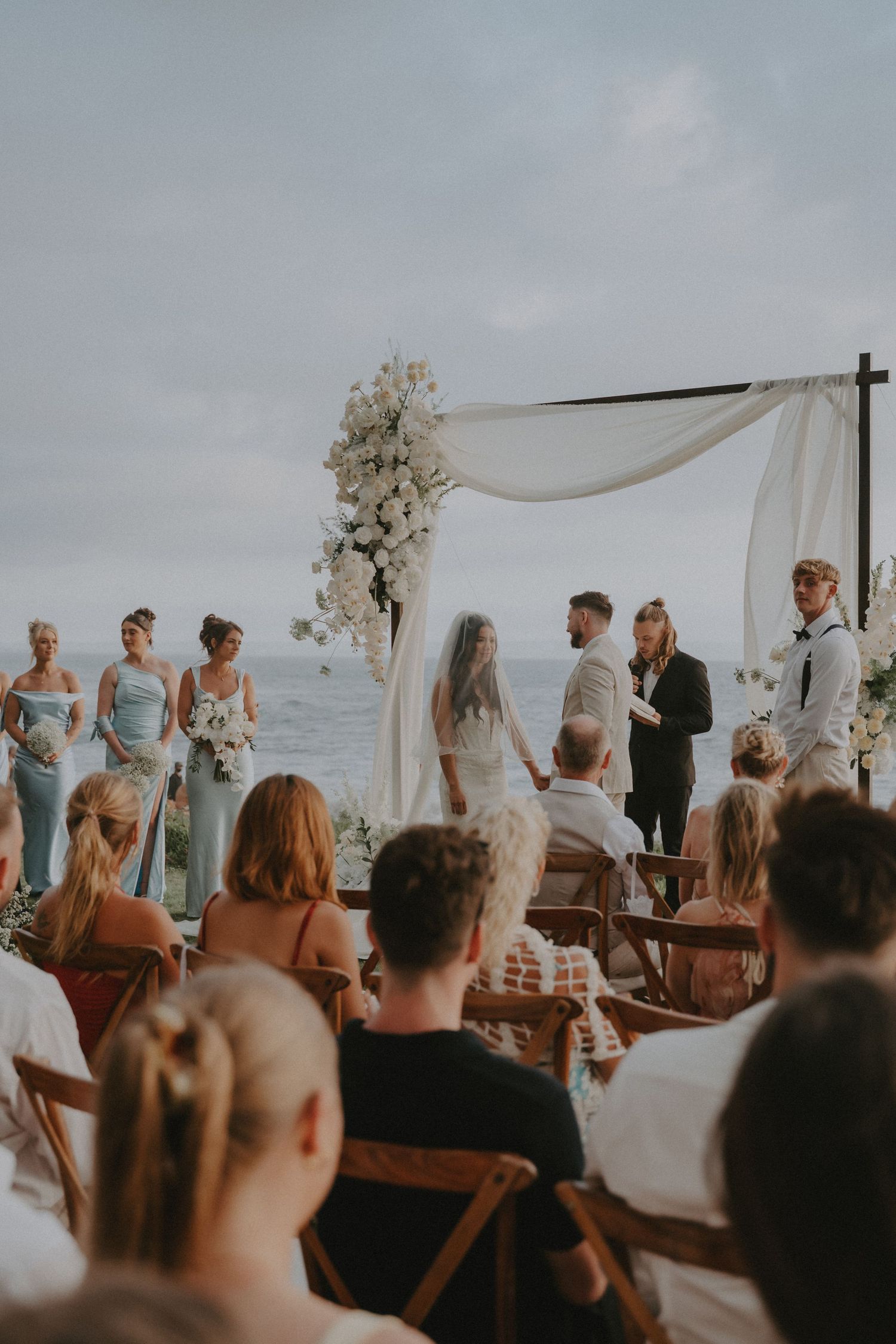 Wedding ceremony guests seated in wooden chairs watch as couple exchanges vows under an outdoor arch at sunset.