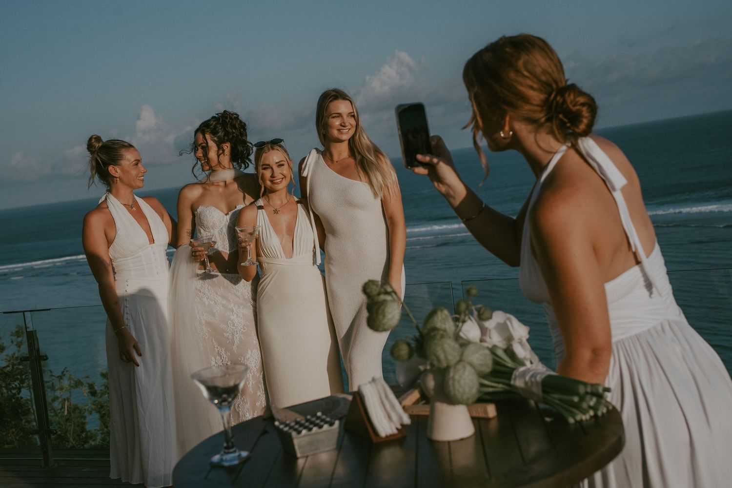 A group in white dresses poses for photos at a scenic ocean overlook during a wedding celebration.
