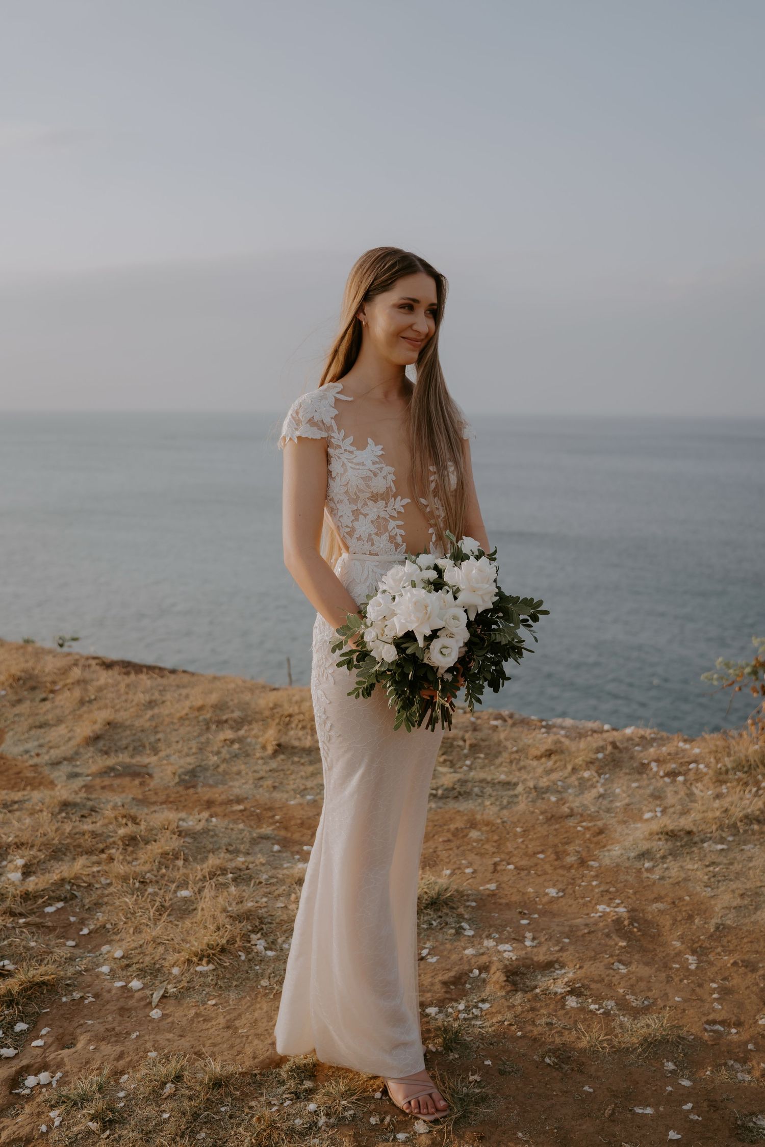 A bride in a delicate lace wedding dress holds a white bouquet while standing on a coastal cliff overlooking the ocean.