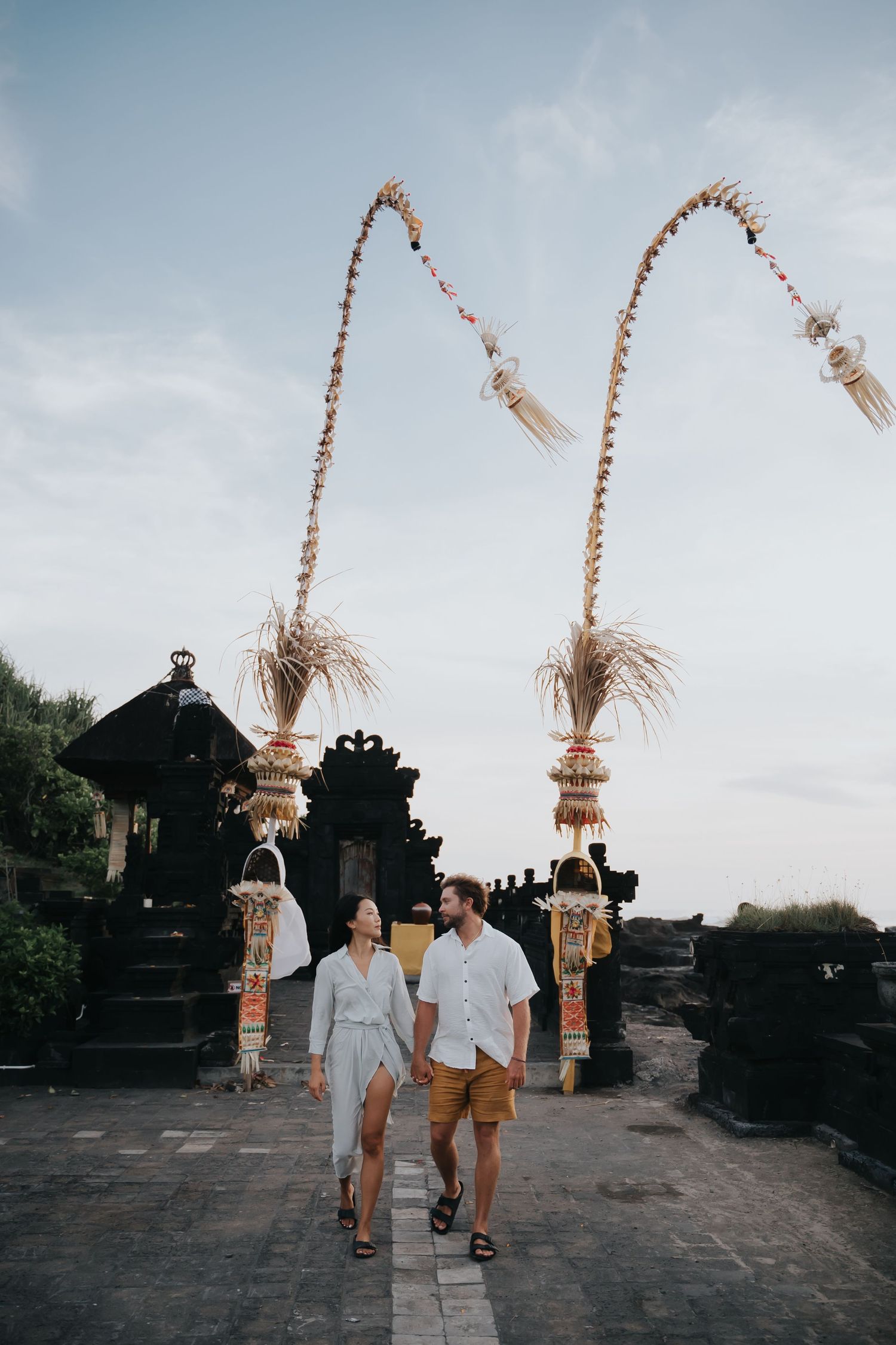 A couple walking through traditional Balinese temple gates with decorative penjor bamboo poles against cloudy sky.