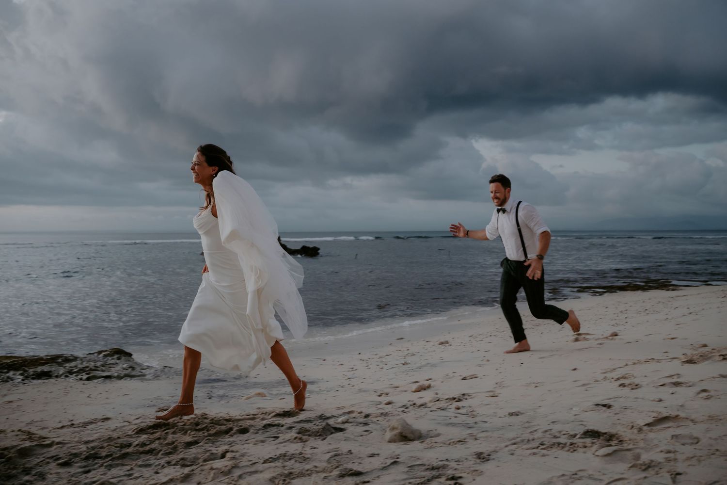 Two people running playfully along a beach under dramatic stormy clouds at dusk.