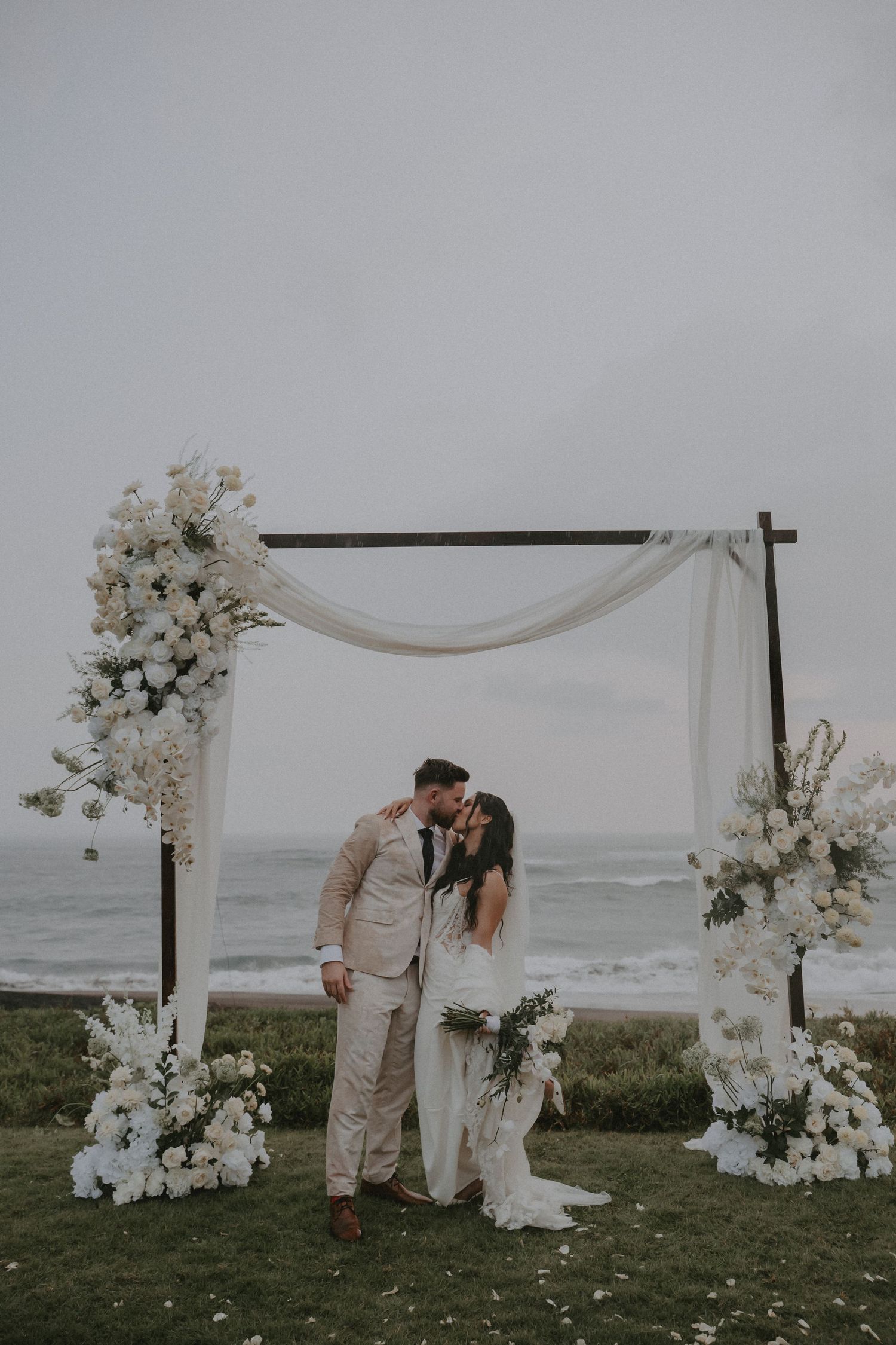 A romantic beach wedding ceremony setup with white floral arrangements and fabric draping against an ocean backdrop.