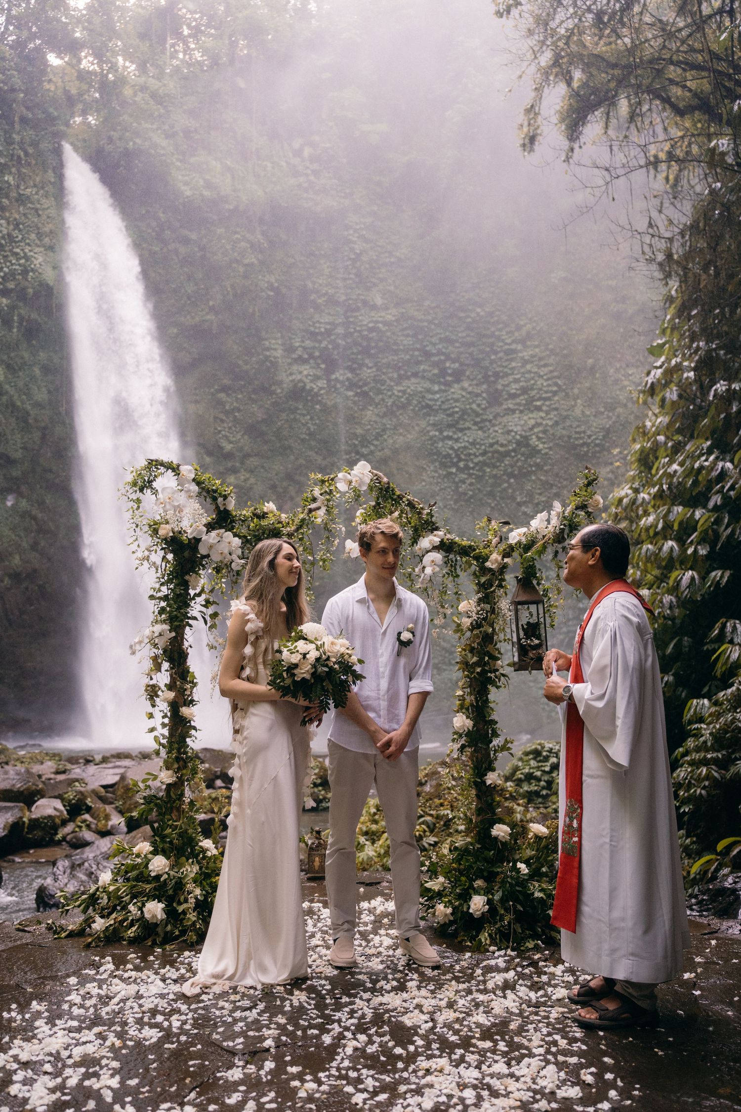 A romantic wedding ceremony takes place in front of a majestic waterfall with a floral arch decorated in white roses and greenery.