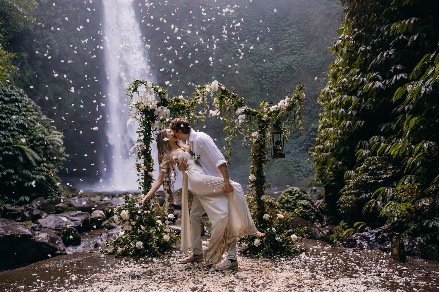 A couple shares a romantic moment under a floral arch with a majestic waterfall cascading in the background.