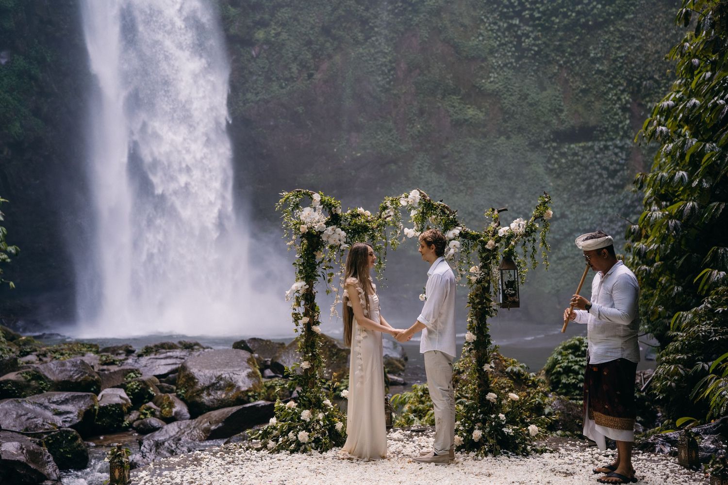 A couple stands in front of a majestic waterfall during their wedding ceremony in a lush tropical setting.