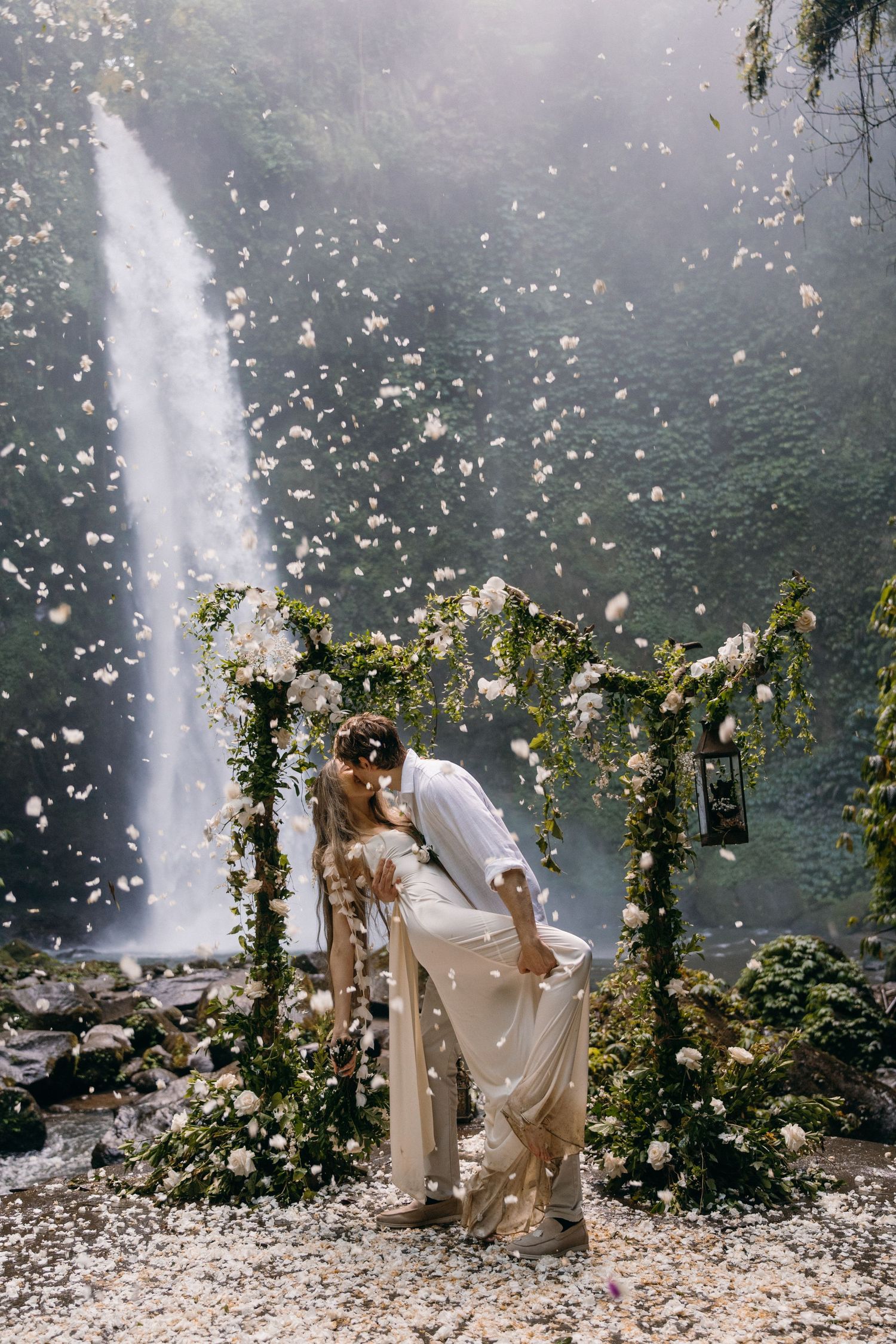 A romantic wedding couple poses in front of a majestic waterfall with floral vine arches and floating petals creating a dreamy scene.