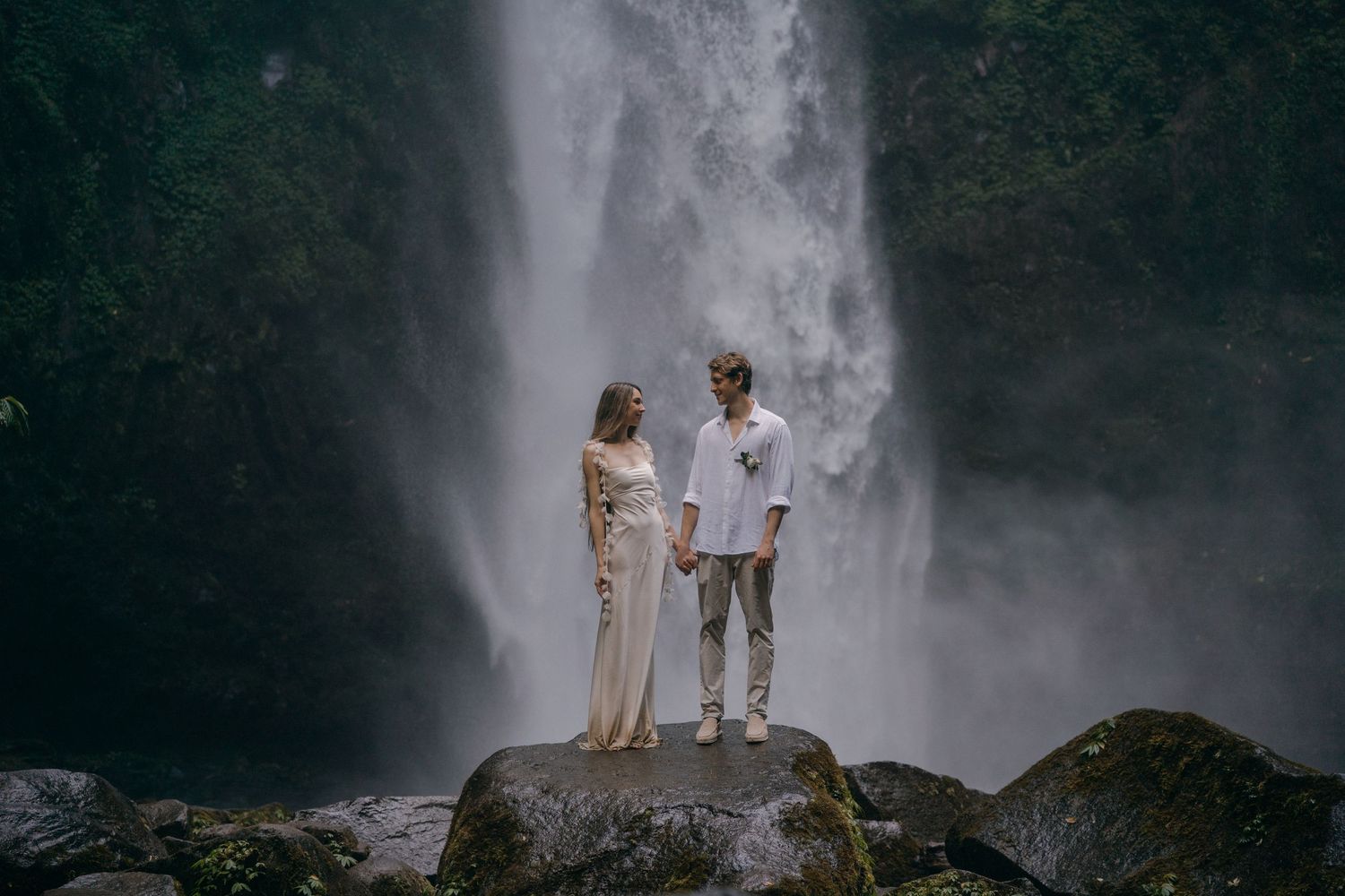 A couple stands together on a rocky outcrop in front of a majestic cascading waterfall.