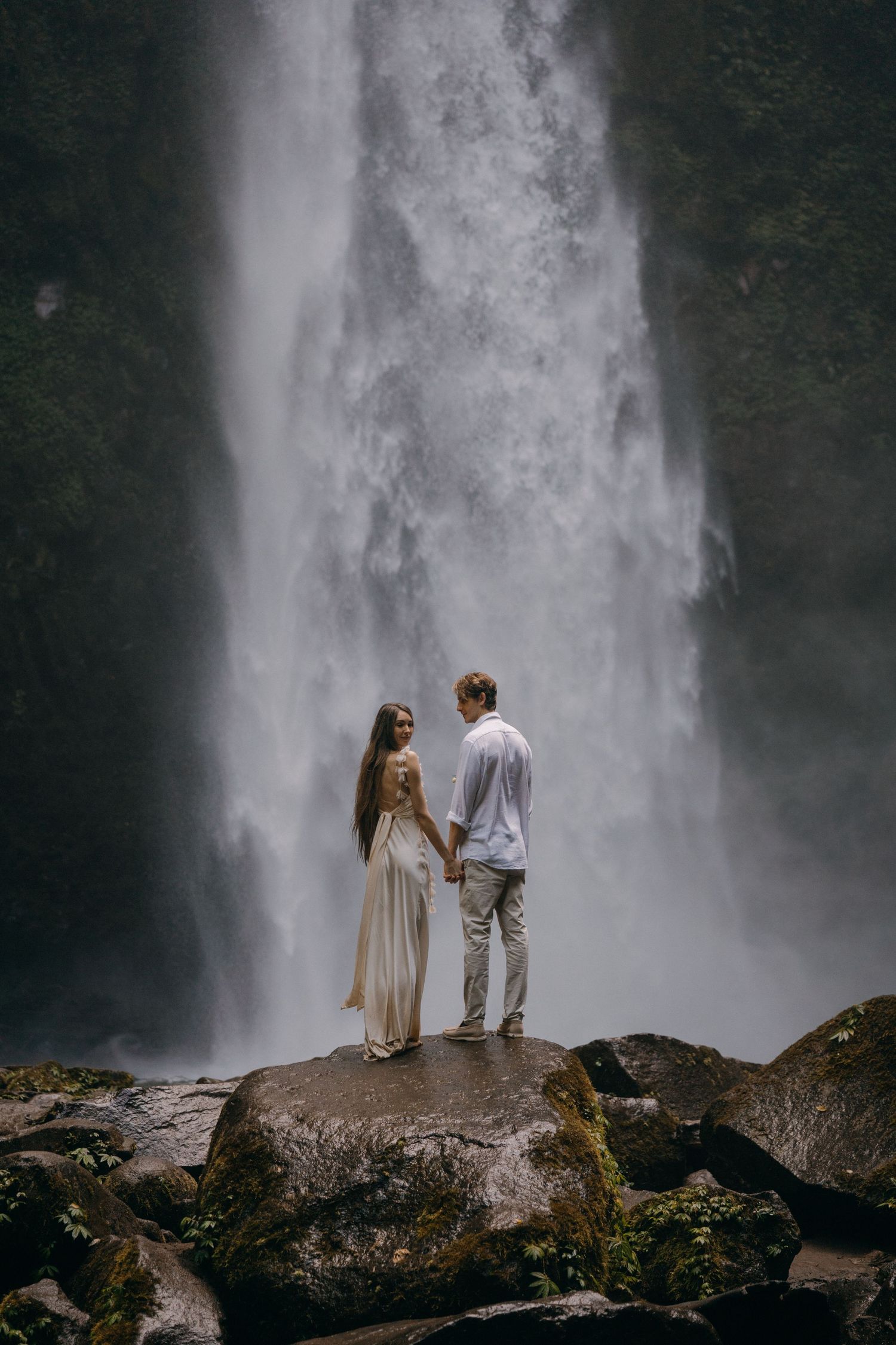A couple stands together on a rocky outcrop before a majestic waterfall in a moody, atmospheric setting.