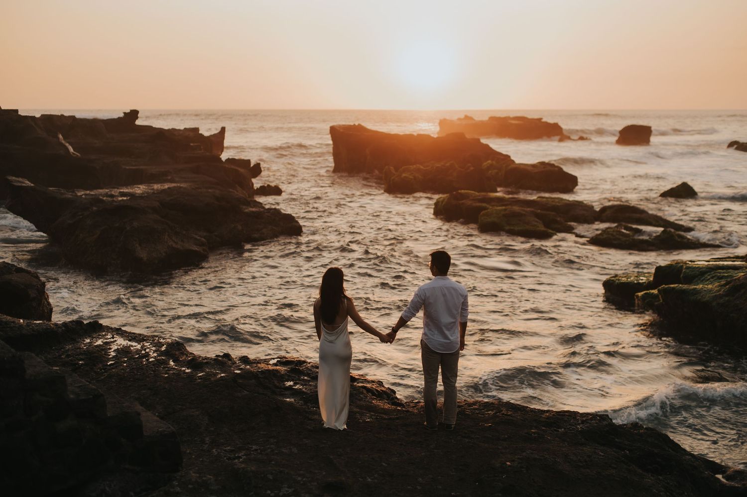 Couple holding hands watching sunset over rocky ocean coastline.