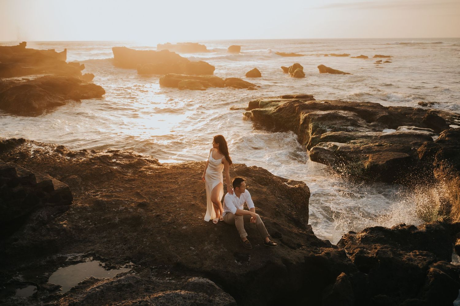 Couple sitting on rocky coastal cliffs at golden sunset with ocean waves crashing behind them.