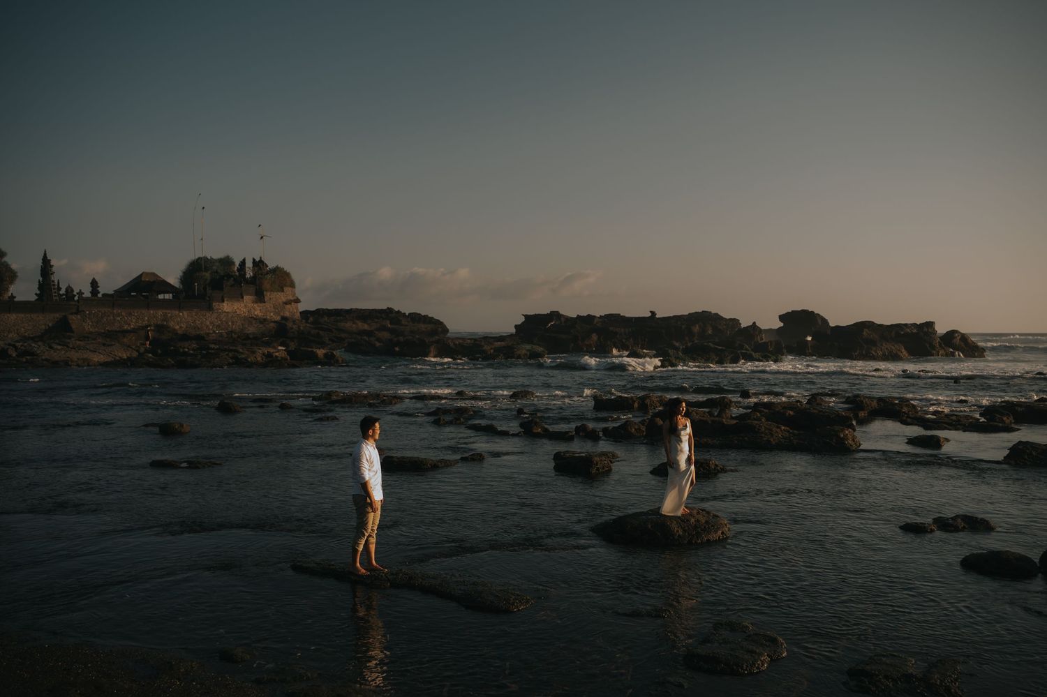 Couple standing on rocks at a dramatic coastal sunset, with ocean waves and ancient temple ruins in the background.