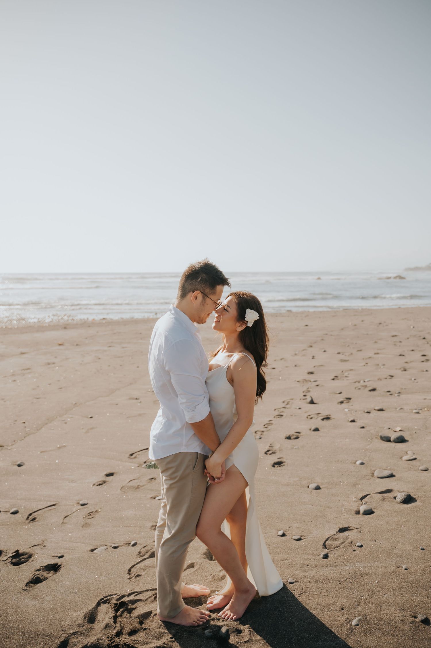Romantic couple embracing on a sandy beach with ocean waves, woman in white dress with flower hair accessory.
