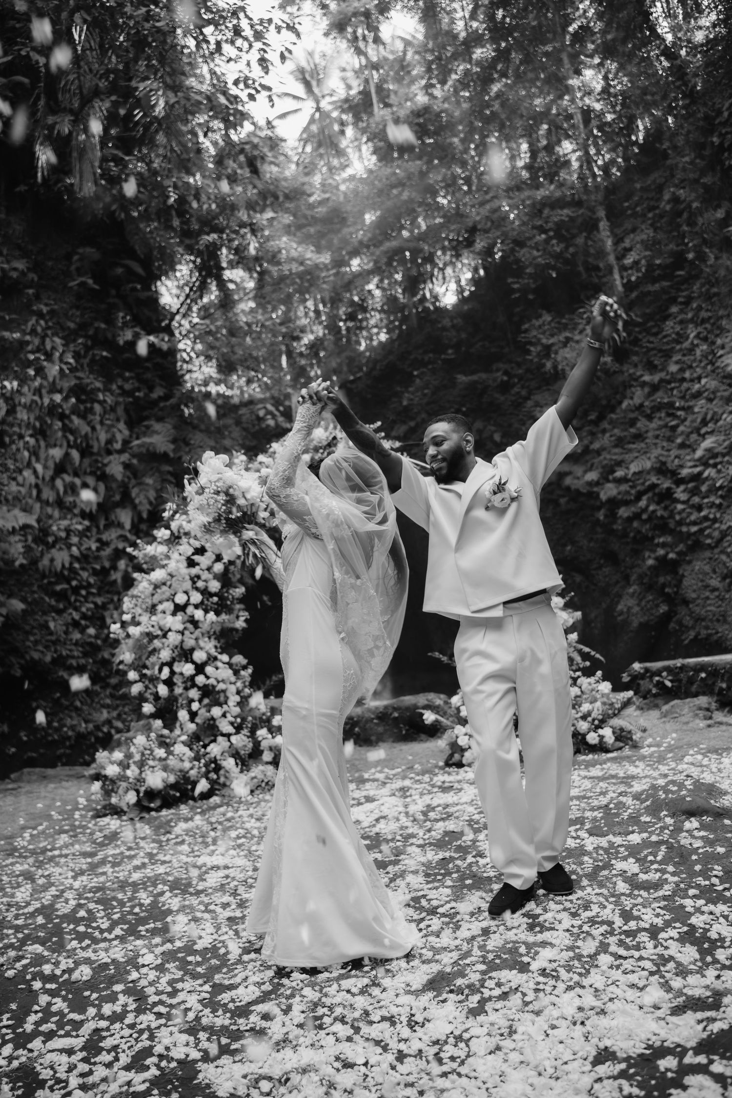 Black and white photo of jubilant newlyweds celebrating with raised arms amid falling petals at waterfall wedding.
