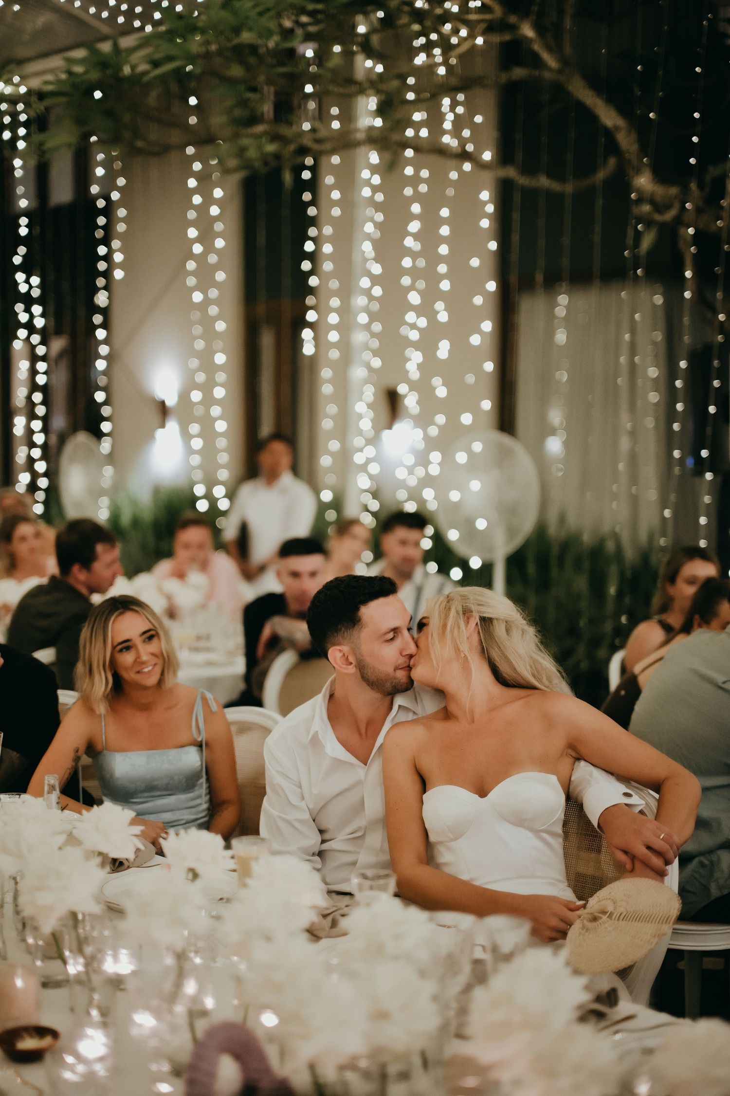 Newlyweds share a romantic kiss at their wedding reception table adorned with white floral centerpieces.