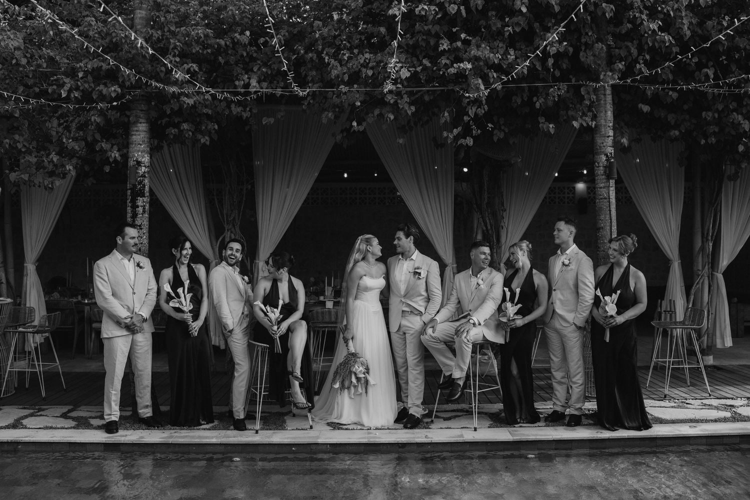 Wedding party poses together under string lights and trees in black and white photo.