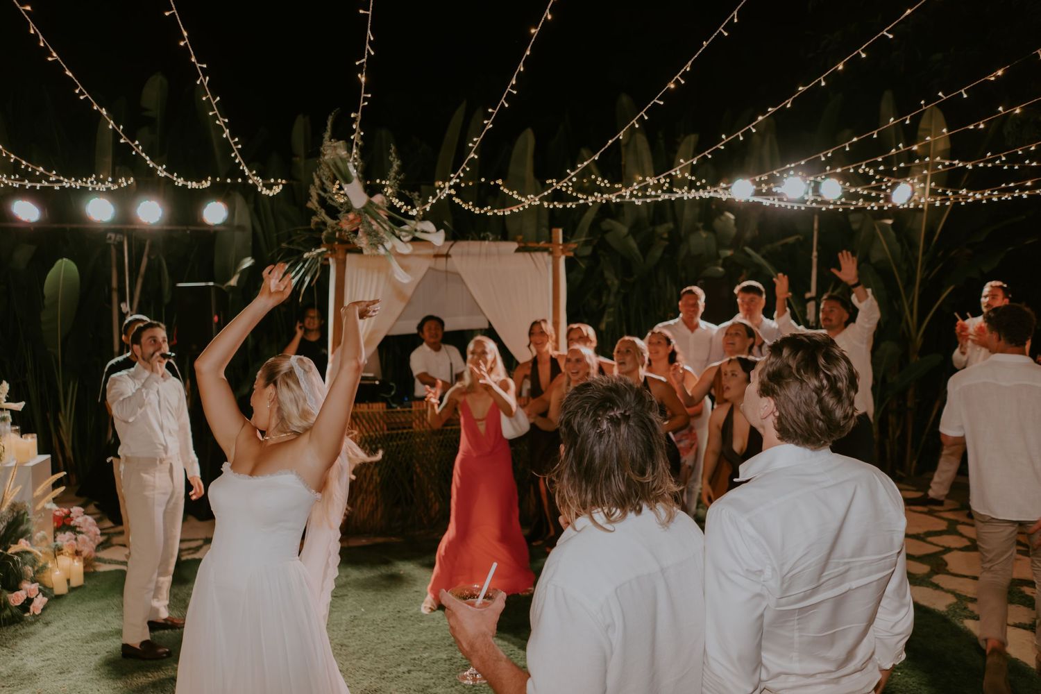 Guests dance under string lights at lively outdoor tropical wedding reception.