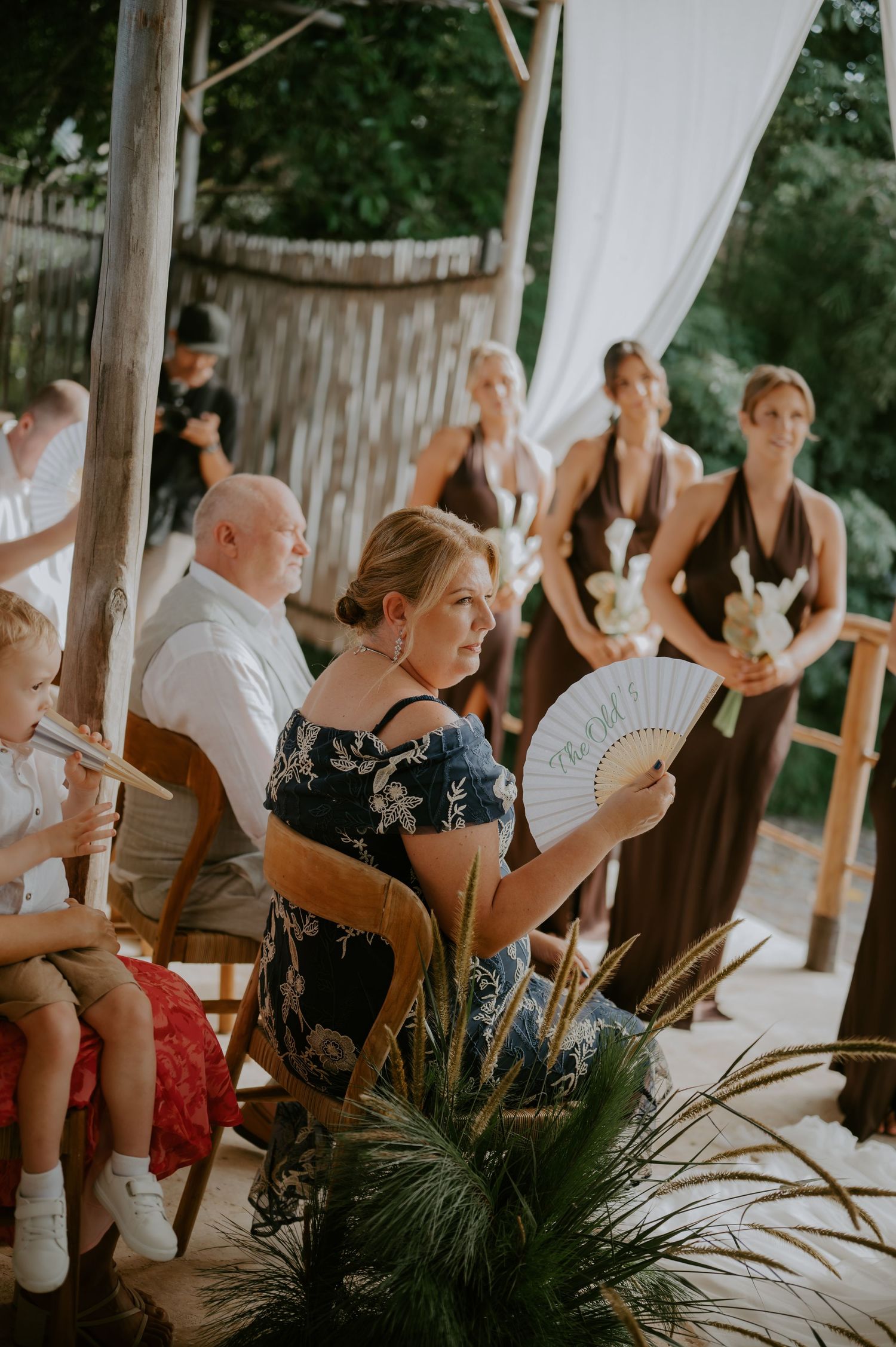 Wedding guest fans herself during outdoor ceremony while bridesmaids in brown dresses stand near floral arch.