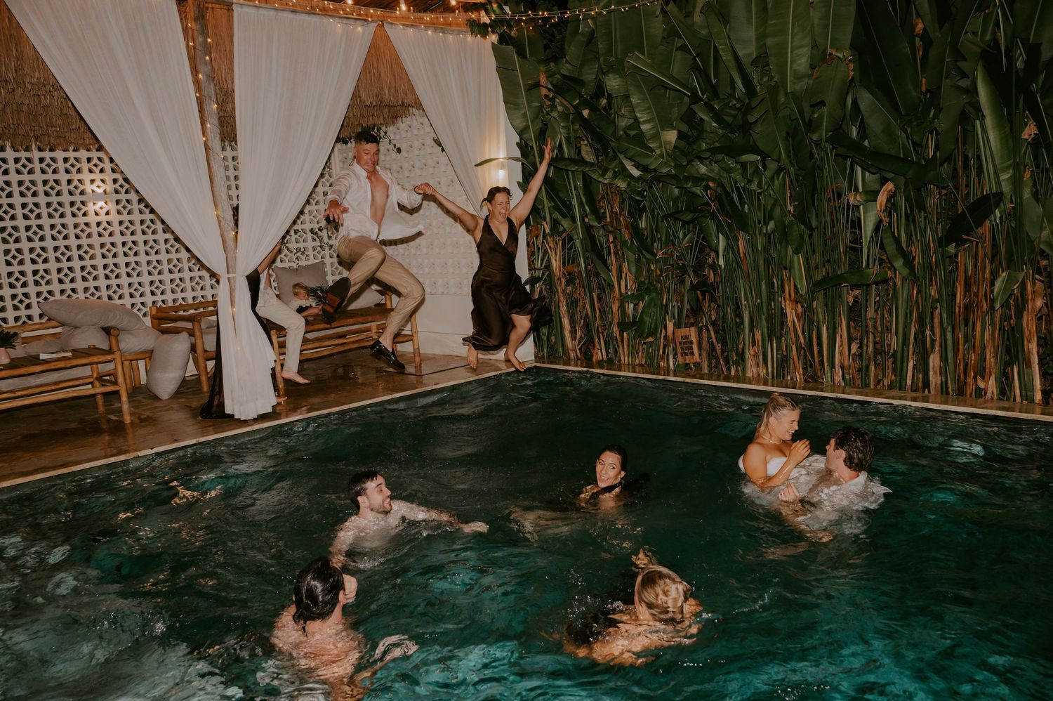 Wedding guests jump into and swim in tropical pool during late-night wedding celebration.