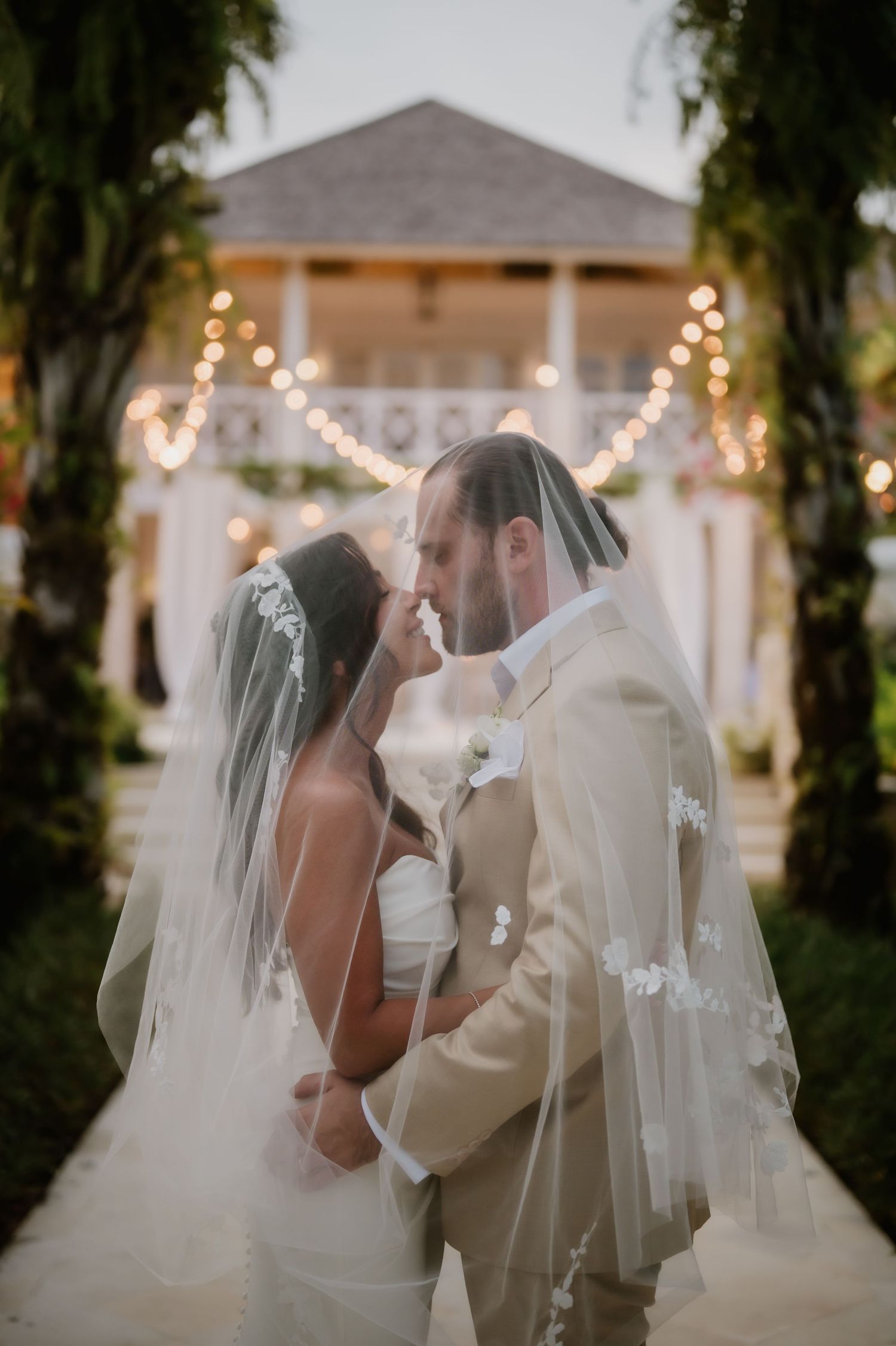 Bride and groom kissing under veil at elegant outdoor wedding venue with string lights.
