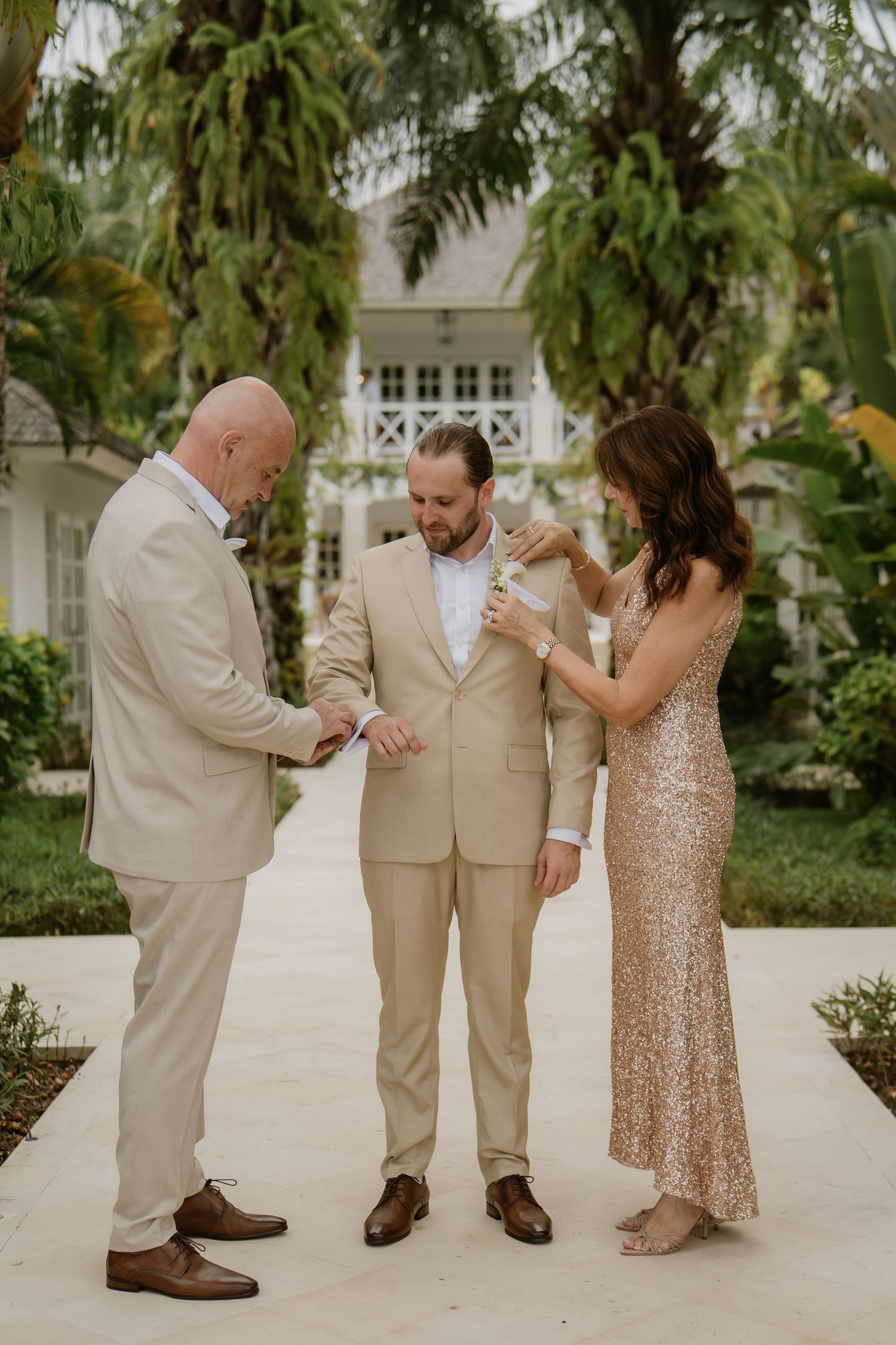 Groom in tan suit being helped with boutonniere by two attendants in a tropical resort setting.