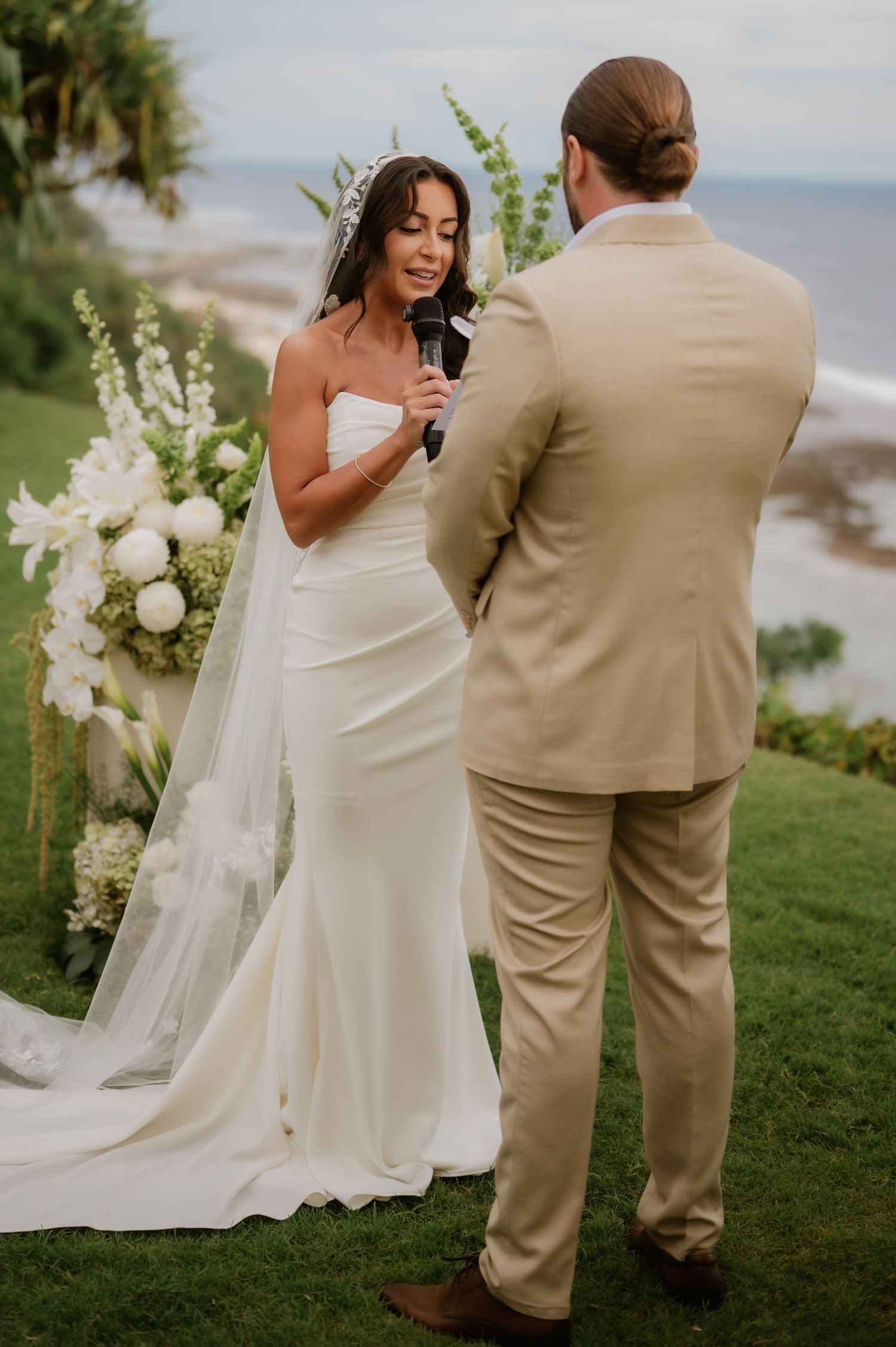 Bride in white gown and veil reciting vows to groom in tan suit at outdoor oceanfront wedding ceremony.