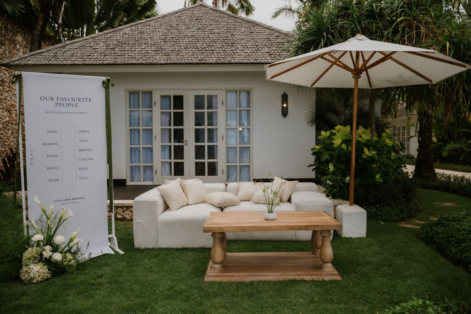 Elegant outdoor wedding lounge with white sofa, wooden coffee table, umbrella, and seating chart sign.
