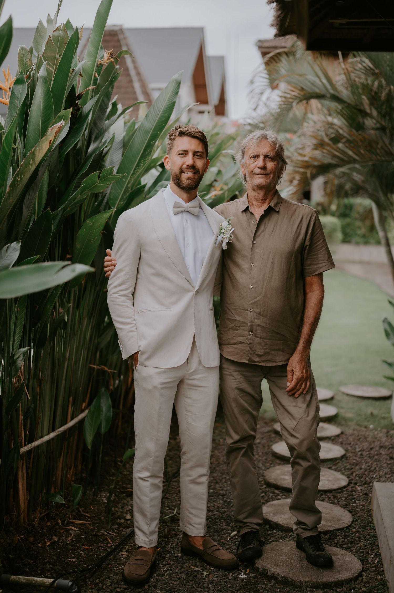 Groom in white suit with boutonniere poses with older man in tropical garden wedding setting.
