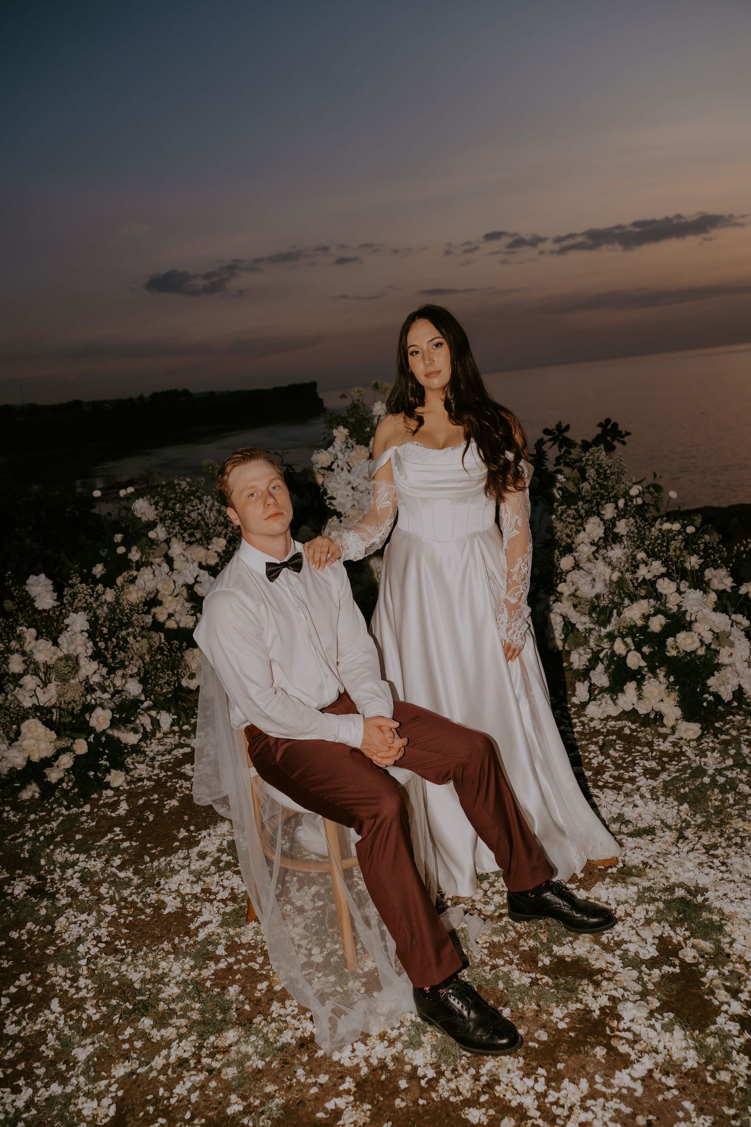 Bride and groom pose at twilight near ocean with white floral arrangements at dusk.