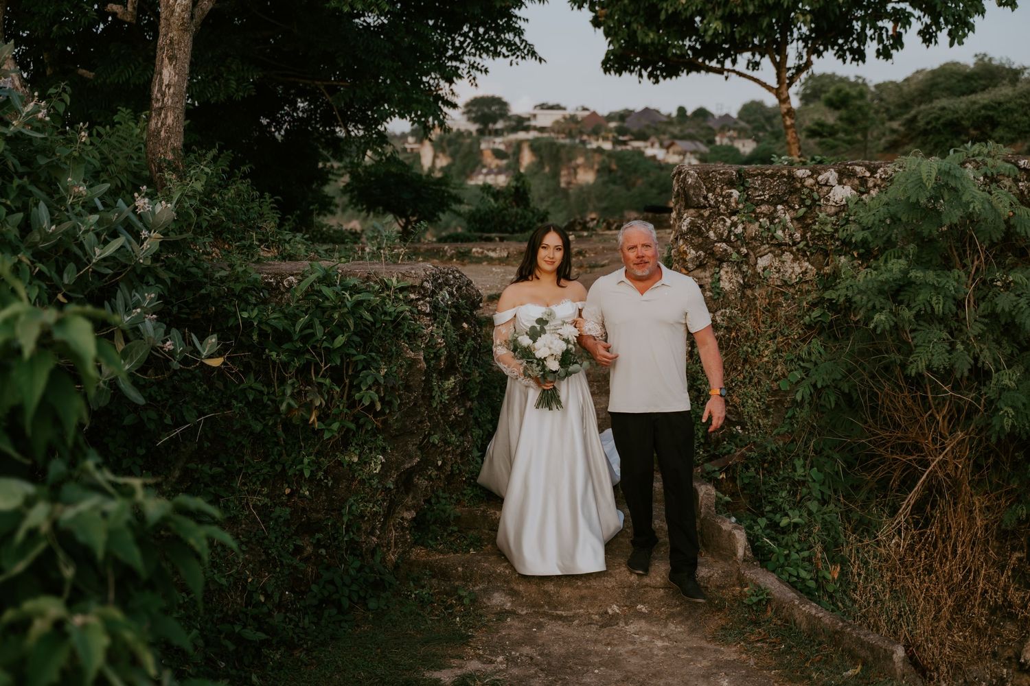 Bride in white gown holding bouquet walks with older man through lush green garden path at outdoor wedding.