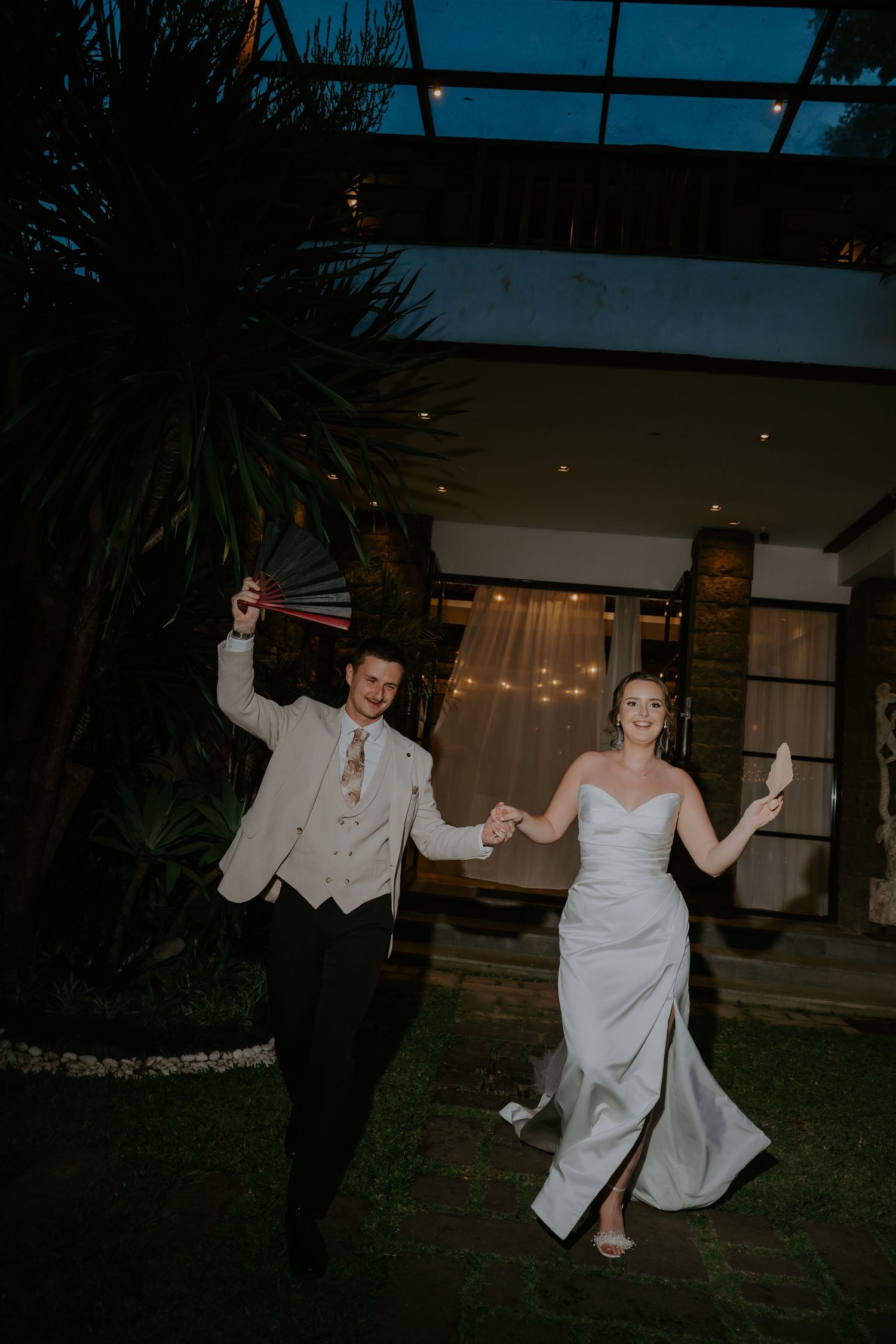 Bride and groom celebrate their grand exit at night, holding sparklers and walking hand-in-hand outside a warmly lit venue.