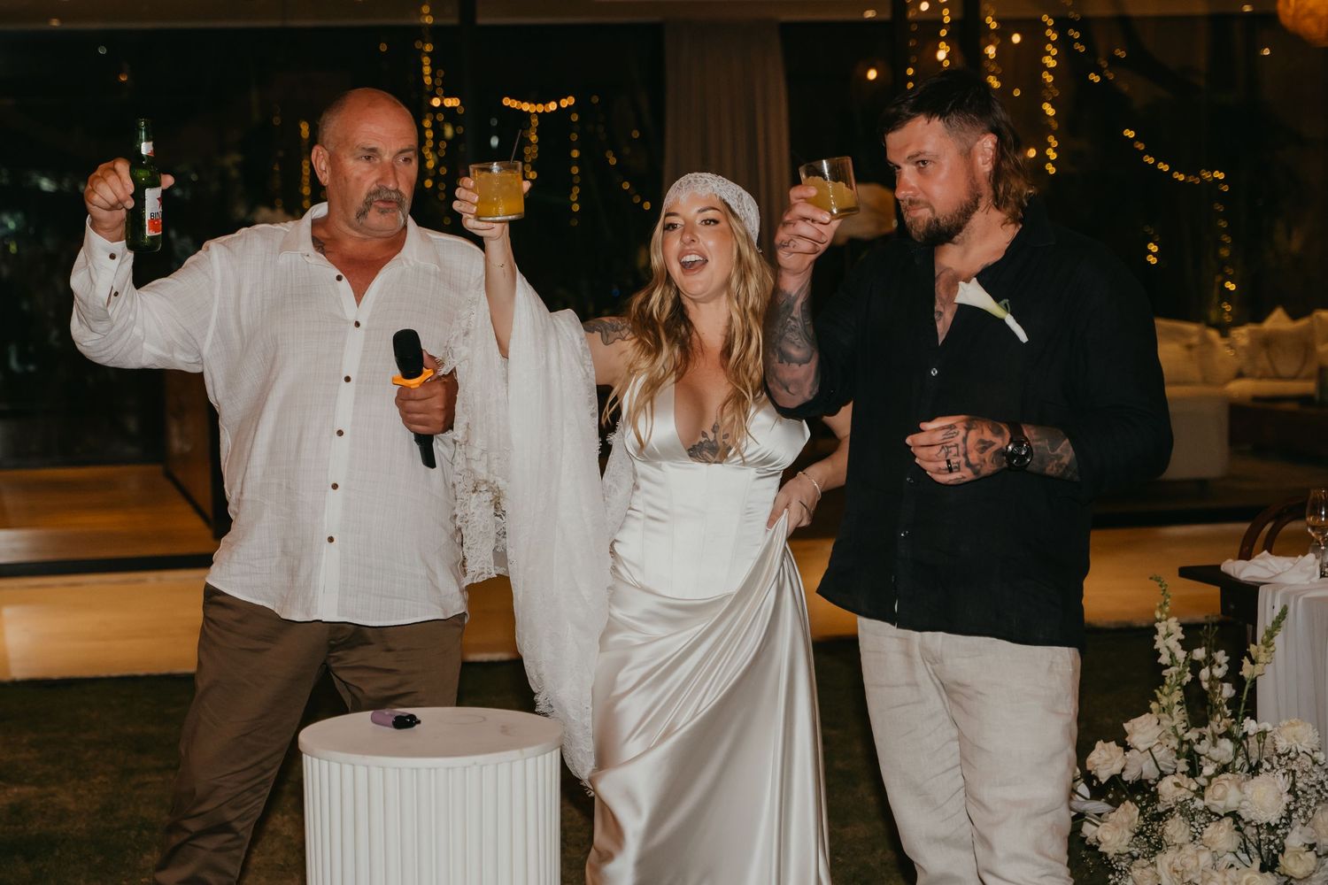 Bride laughs with groom and father during wedding speech toast at elegant floral reception venue.