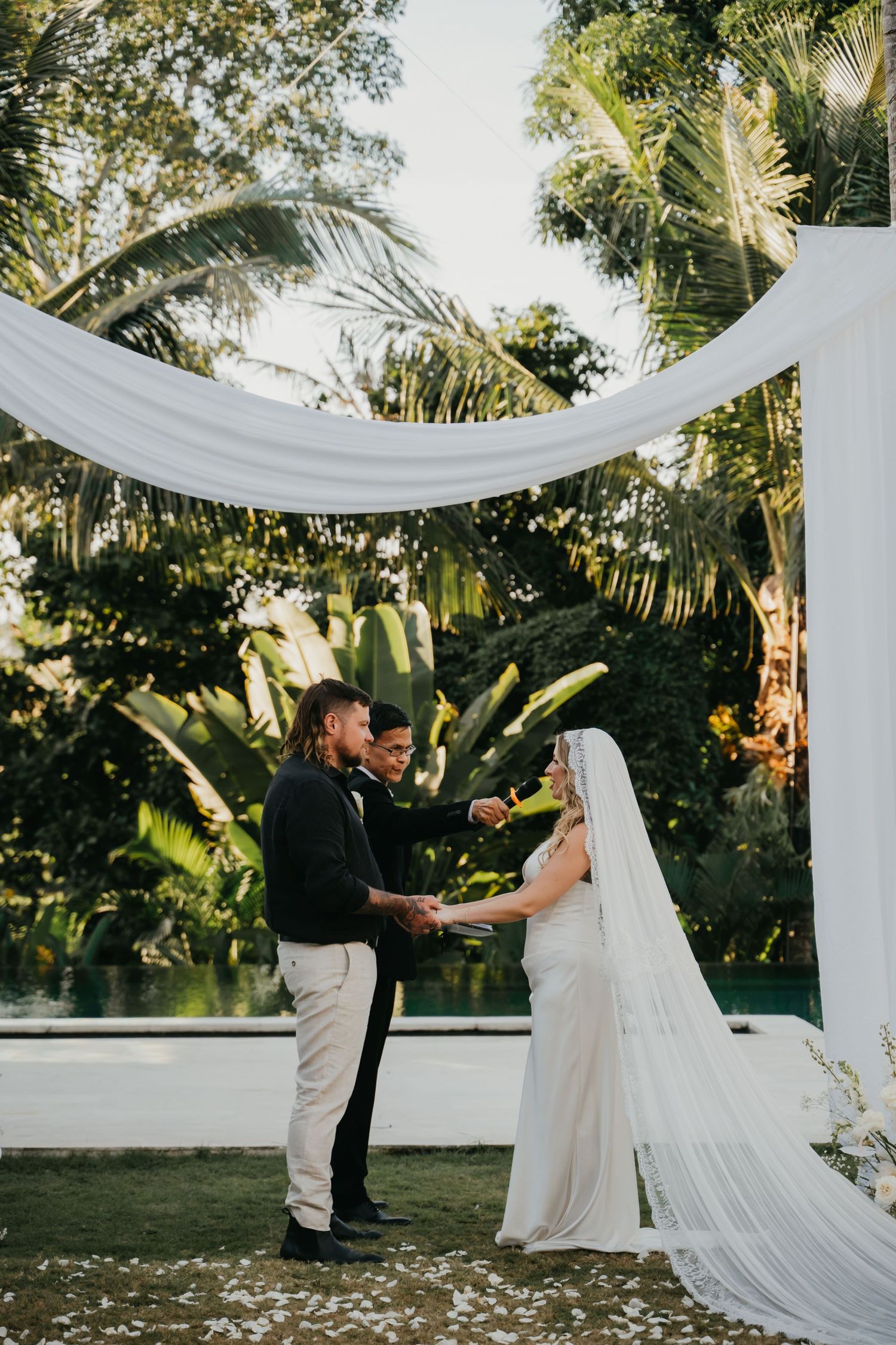 Bride and groom exchange vows under a white draped arch surrounded by lush tropical greenery at an outdoor wedding ceremony.