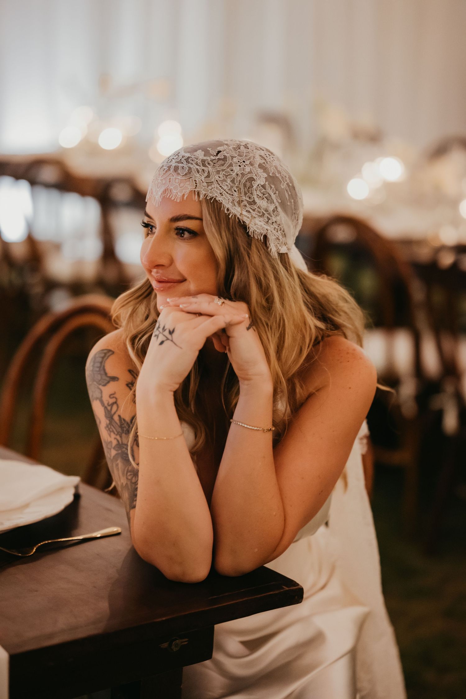 Tattooed bride in satin dress and lace bridal cap poses thoughtfully at reception table.