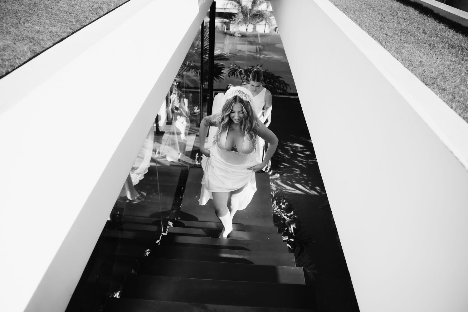 Black and white photo of bride in wedding dress descending modern staircase with water feature.