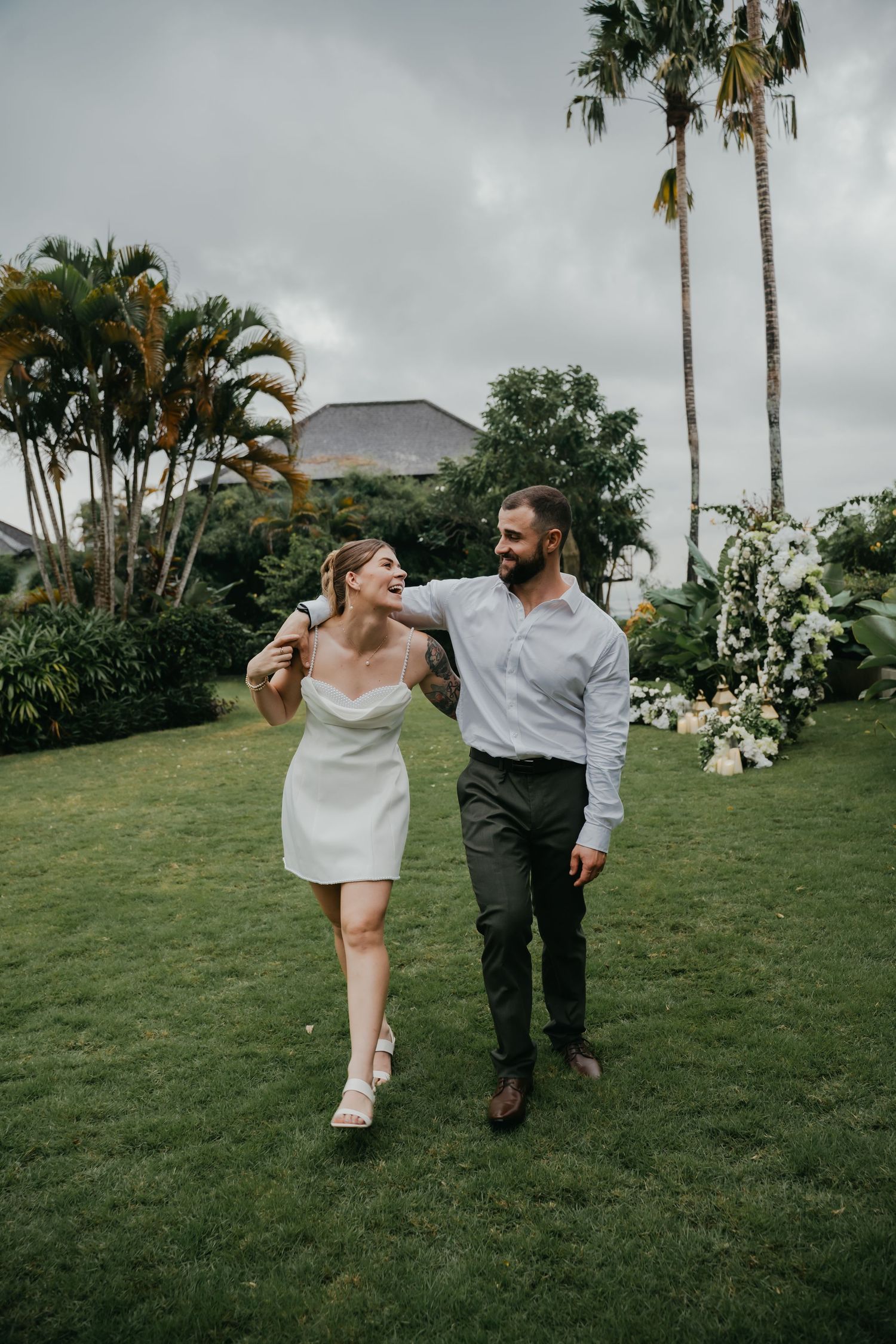 Laughing newlywed couple walks together across green lawn in casual wedding attire after ceremony.