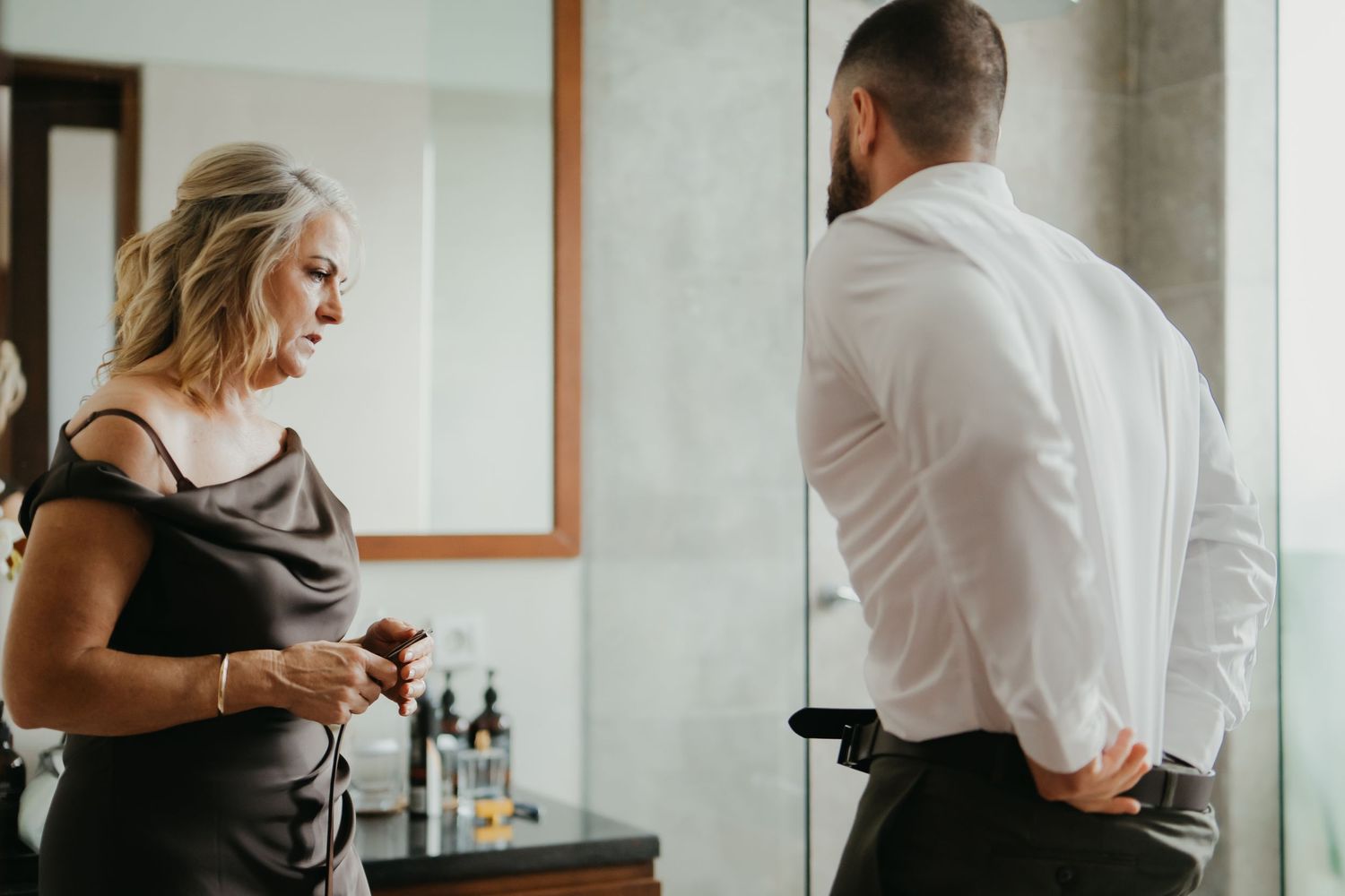 Woman in black dress helps groom button his white dress shirt in elegant hotel bathroom.
