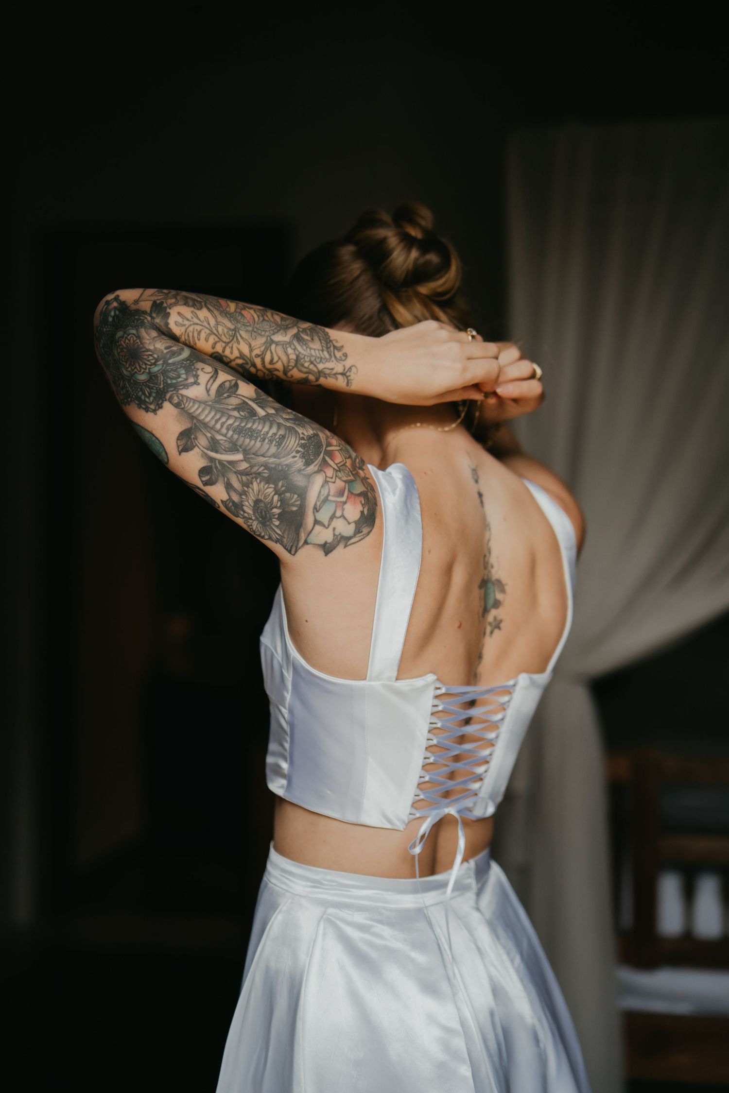 Tattooed bride in a white corset wedding top adjusting her hair, showing intricate back tattoos.