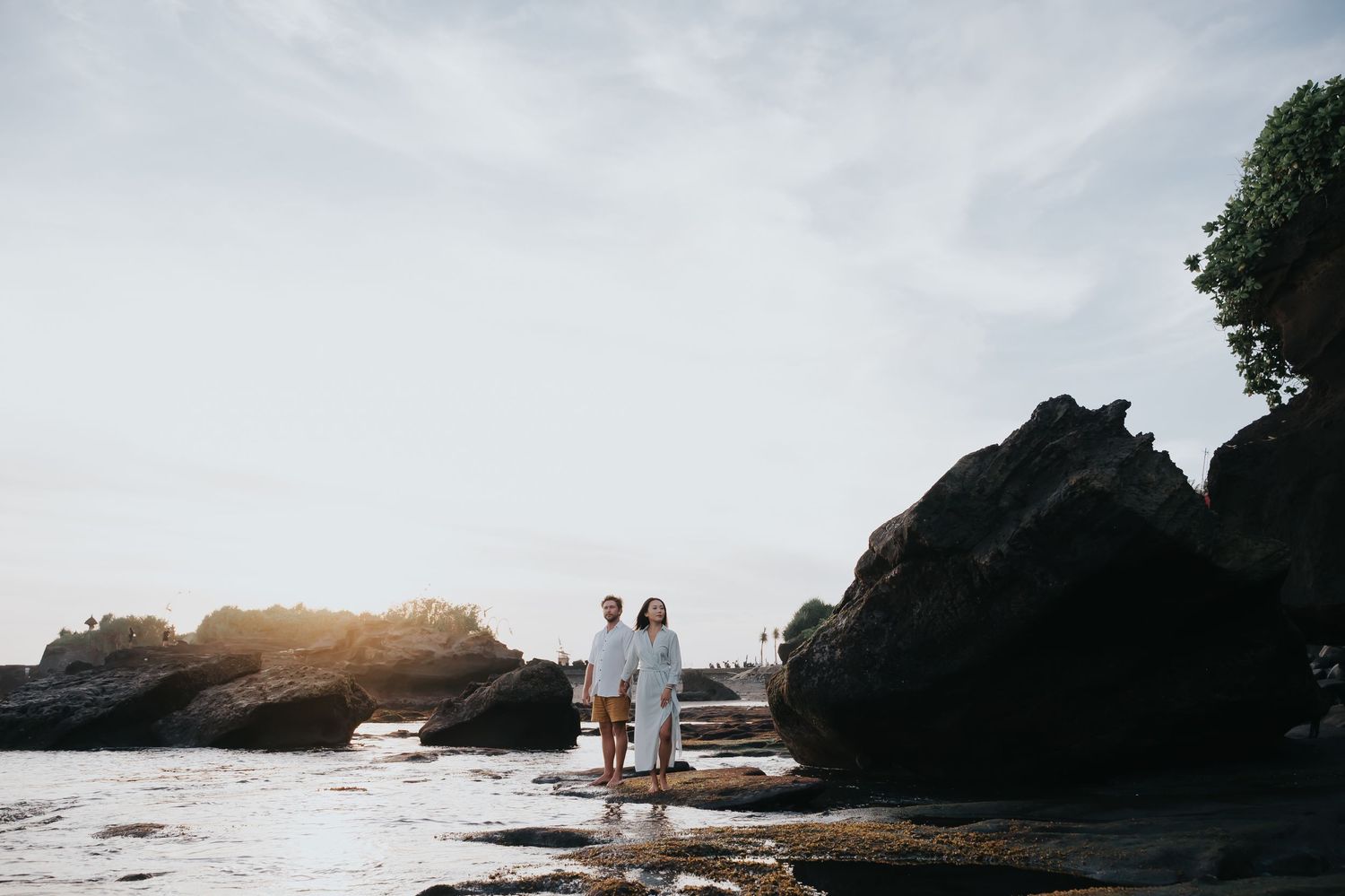 Silhouetted figures stand on rocky coastal cliffs at sunset with ocean waves crashing against the shore.