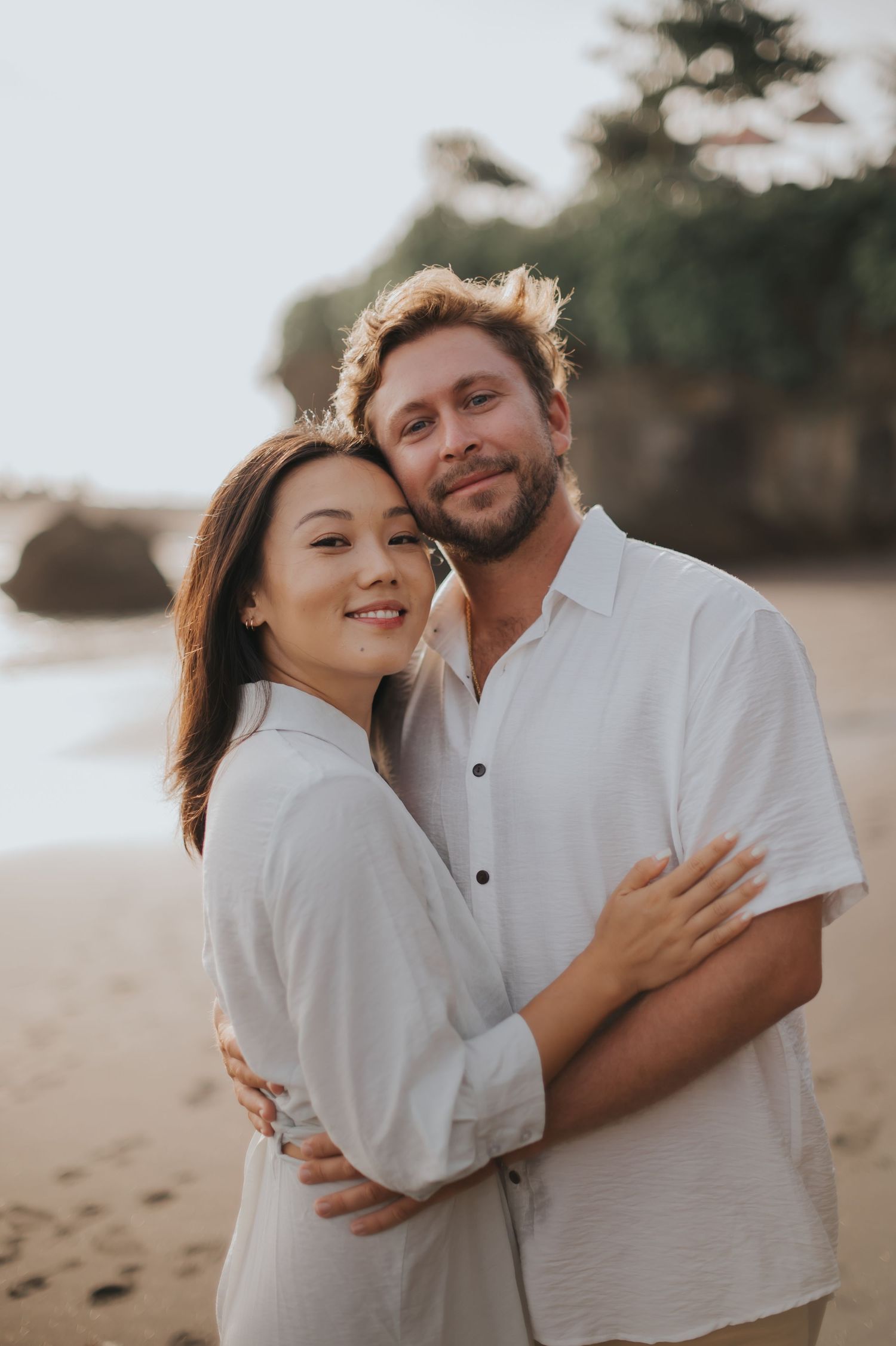 A couple wearing white shirts embrace on a beach at sunset, creating a romantic golden-hour portrait.