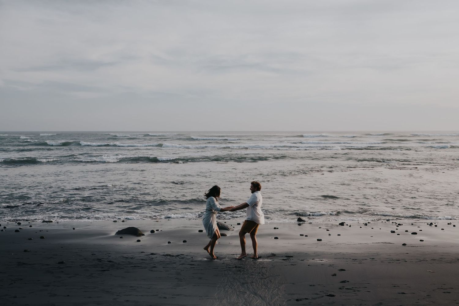 Silhouetted figures explore a misty beach at low tide during sunset with waves crashing in the background.
