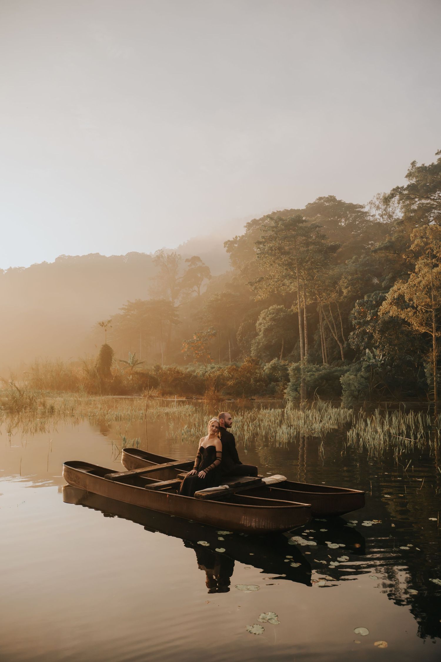 Moody lake landscape with wooden boats drifting through morning fog near wooded shoreline.