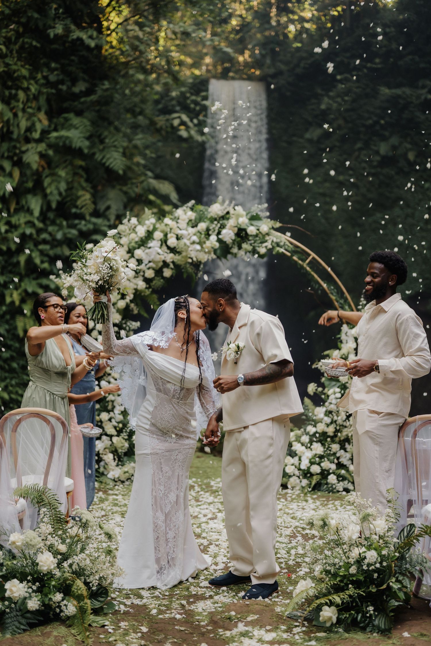 Joyful newlyweds kiss as guests toss white flower petals at outdoor tropical waterfall wedding ceremony.