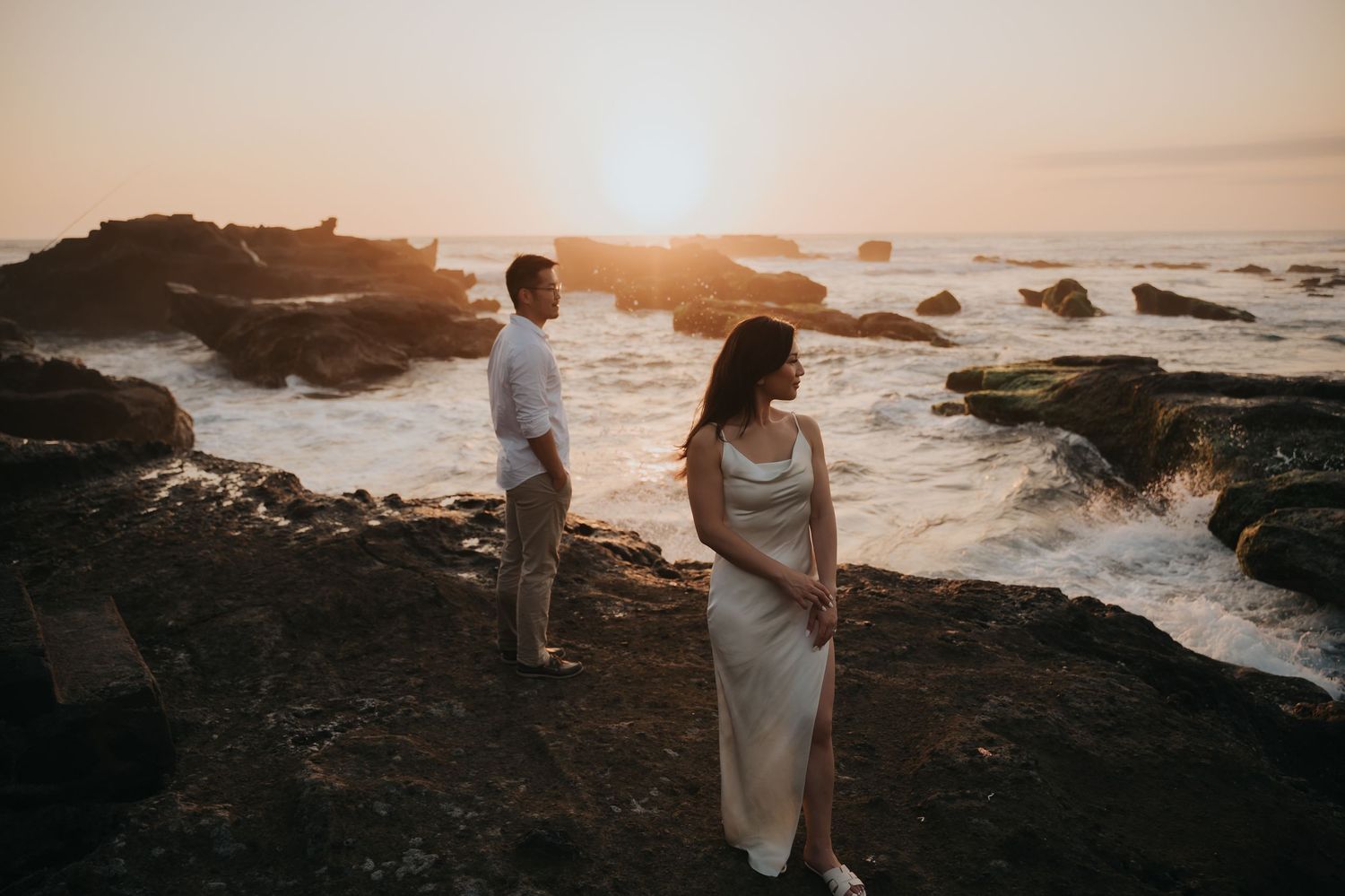 Couple standing on rocky coastal cliffs at golden sunset, waves crashing behind them, woman in elegant white gown.