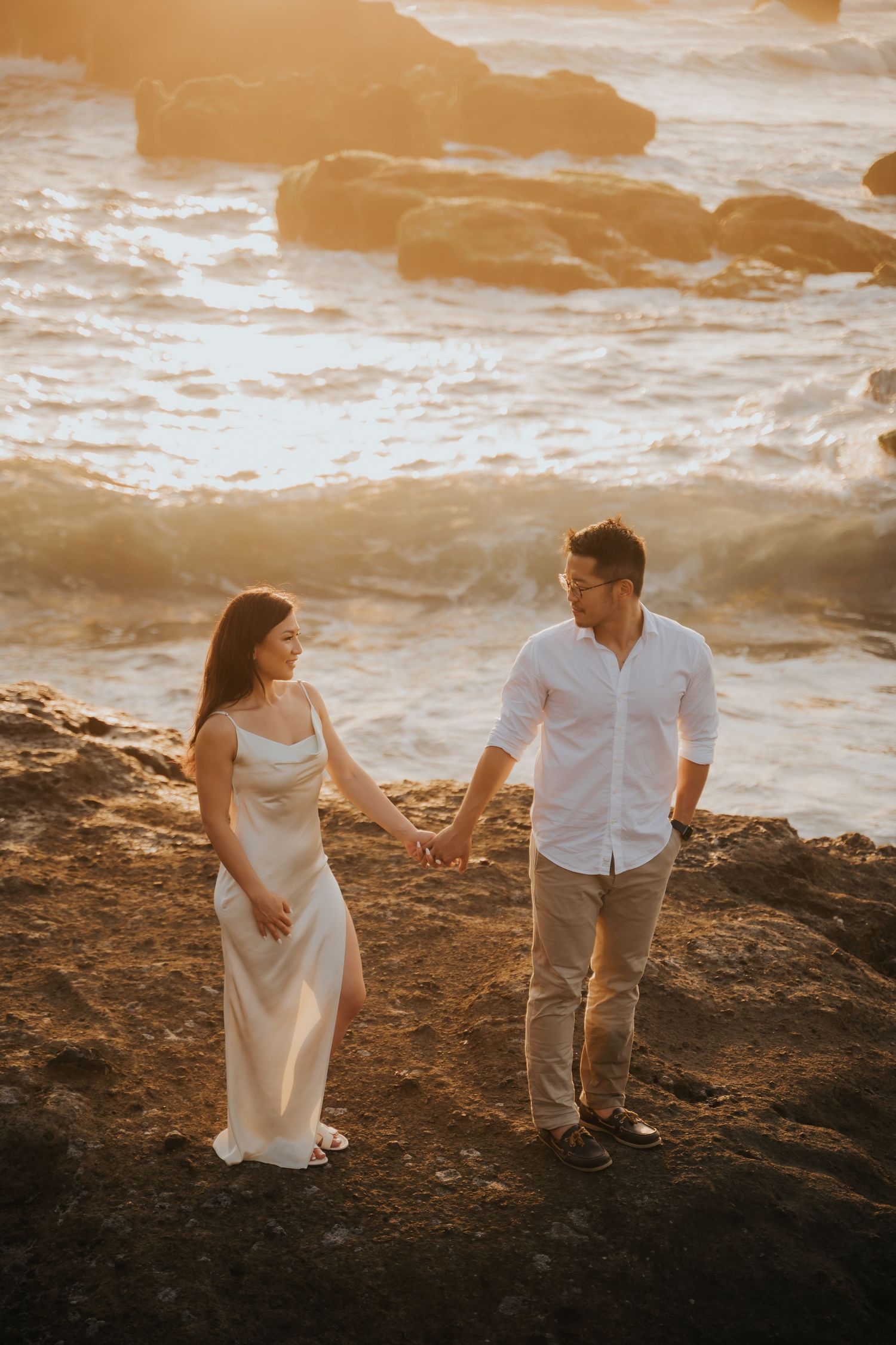 Couple holding hands on rocky coastal cliffs at golden sunset, woman in white dress, man in white shirt.