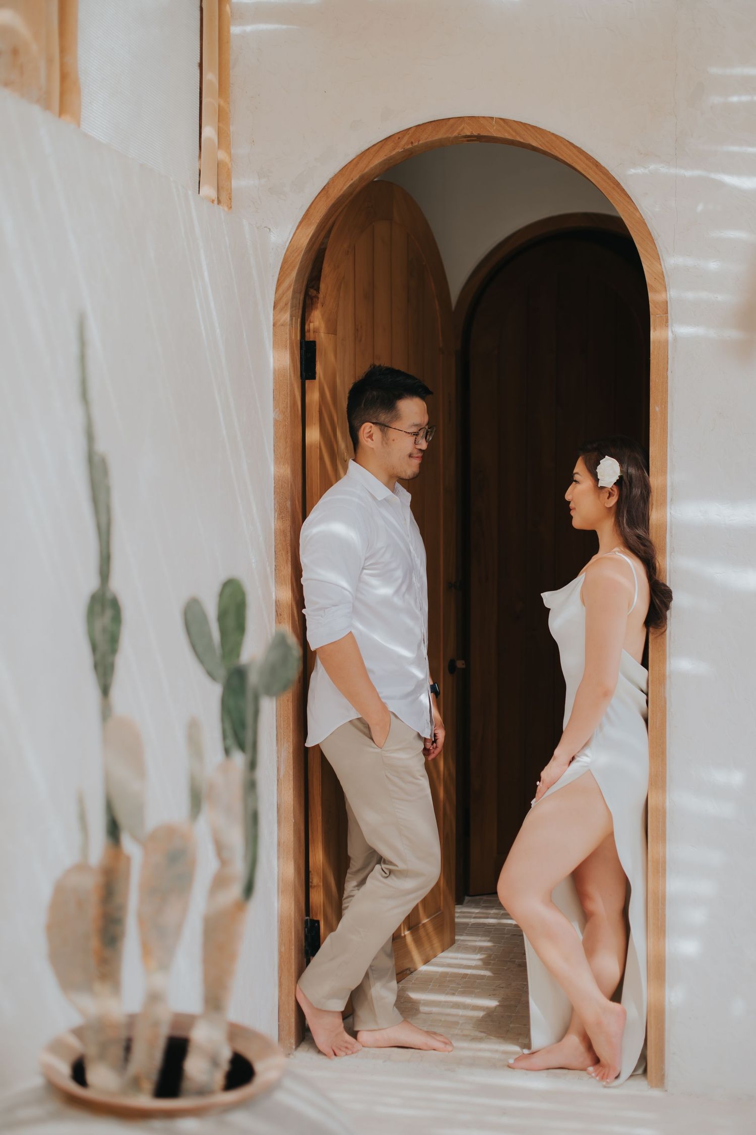Couple in white outfits standing in an arched wooden doorway, sharing a romantic moment in a bright, airy space.
