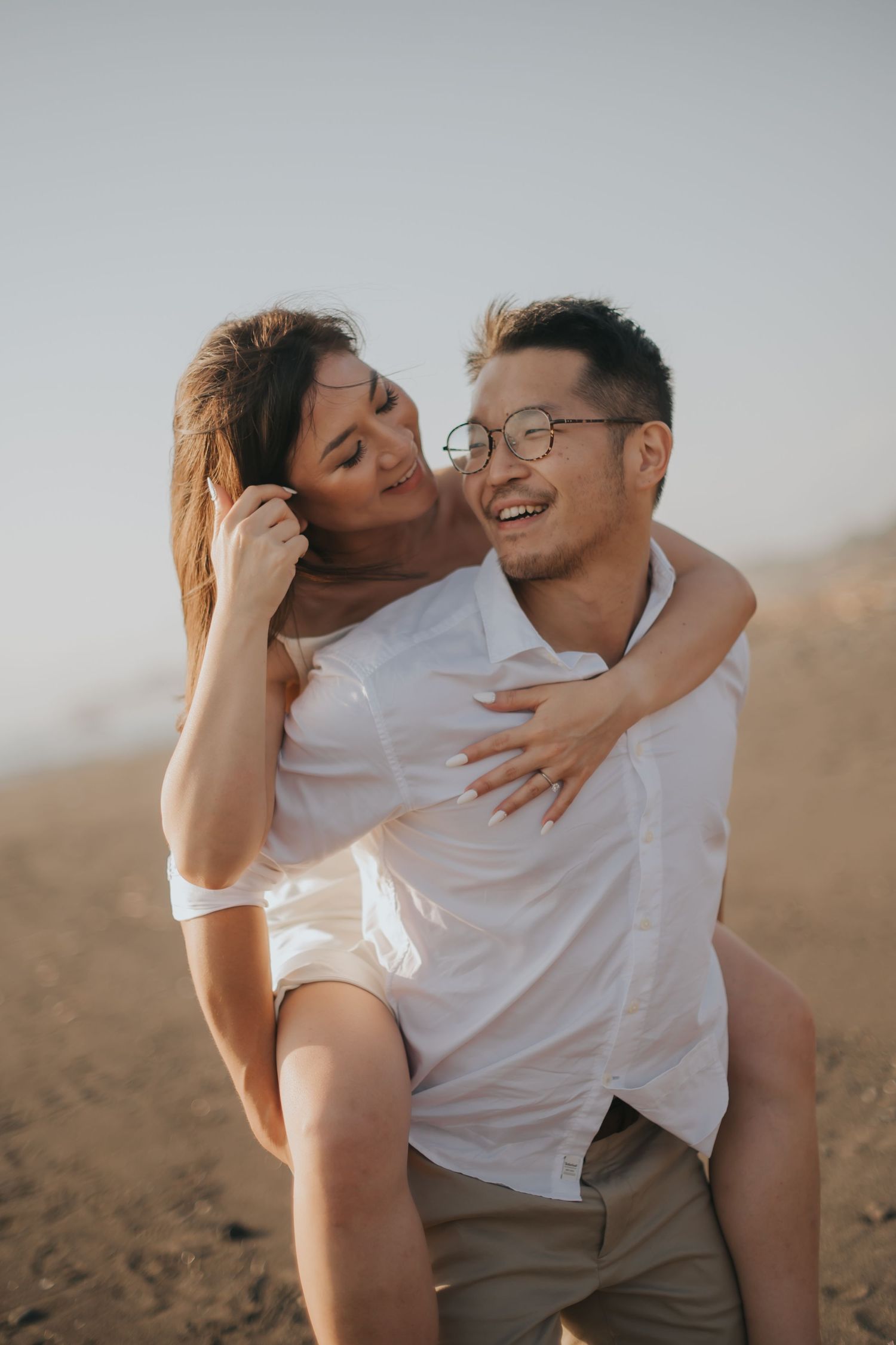 A happy couple sharing a piggyback ride on a sandy beach, both wearing white, smiling joyfully.