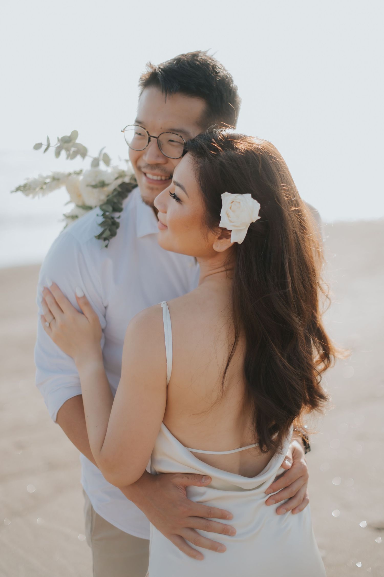 Couple embracing on beach, woman in white dress with flower in hair, man in white shirt holding bouquet.