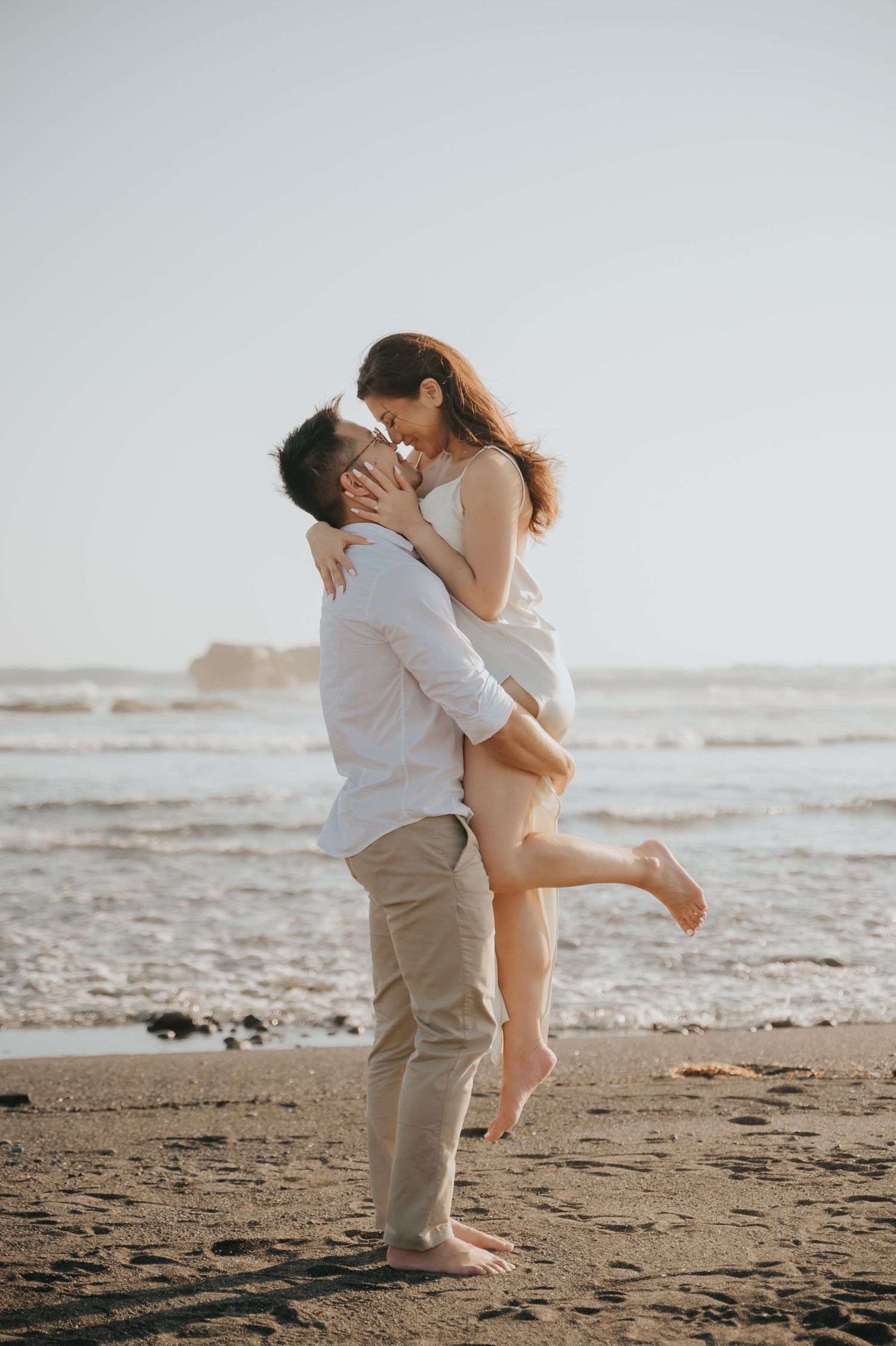 A romantic couple sharing a kiss on a sandy beach at sunset, with the man lifting the woman in a white dress.